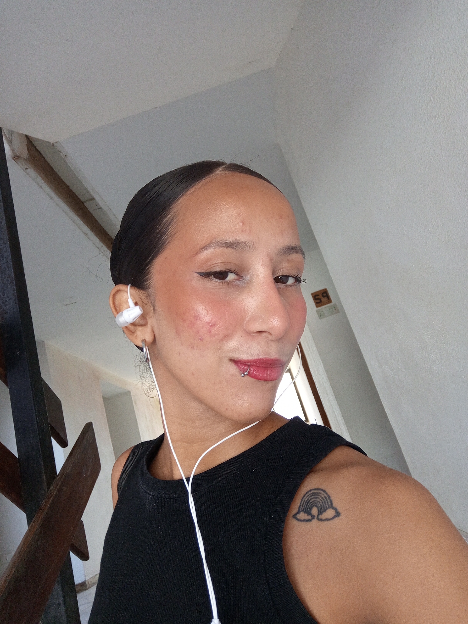 A medium shot depicts a light-skinned person with slicked-back dark hair, wearing a black tank top, and white earbuds. They have a lip piercing on the left side of their lower lip and are wearing large hoop earrings. A tattoo of a rainbow with clouds is visible on their left upper arm. The person is looking at the camera with a slight smile. The background includes a staircase with wooden steps and a white wall. A small, dark object with the number "59" is mounted on the wall. The location is Porlamar, Venezuela, but further details regarding the specific location within the city cannot be determined from the image.