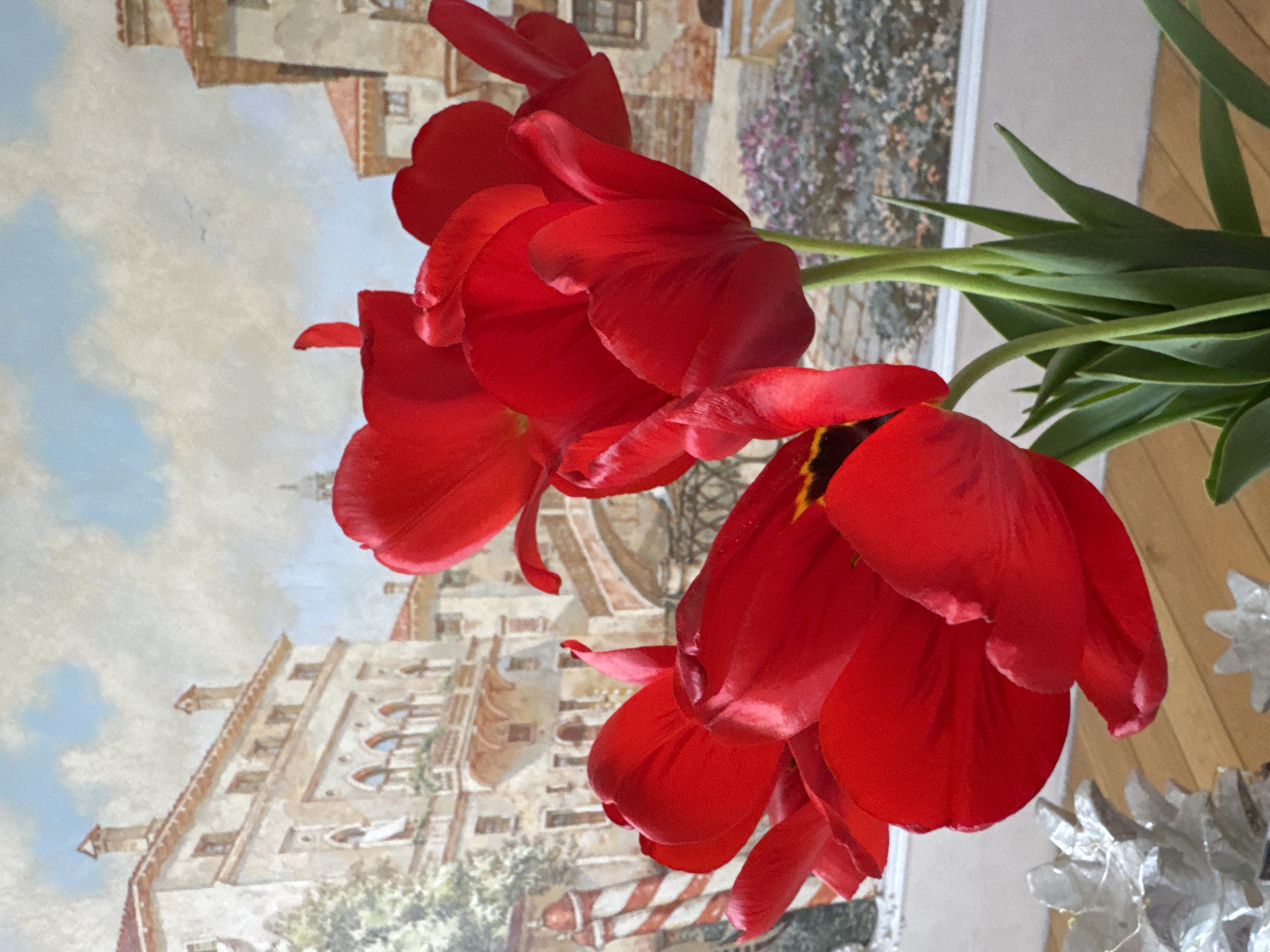 A bouquet of red tulips stands in front of a mural depicting what appears to be a Venice cityscape. The mural shows buildings with arched windows, likely inspired by Venetian architecture. The sky in the mural is a pale blue with scattered clouds. In the foreground, several fully bloomed red tulips with green stems fill the frame. Beside the bouquet is a glass object with a snowflake pattern. The exact location in Saint Petersburg, Russia cannot be determined based on the image provided.
