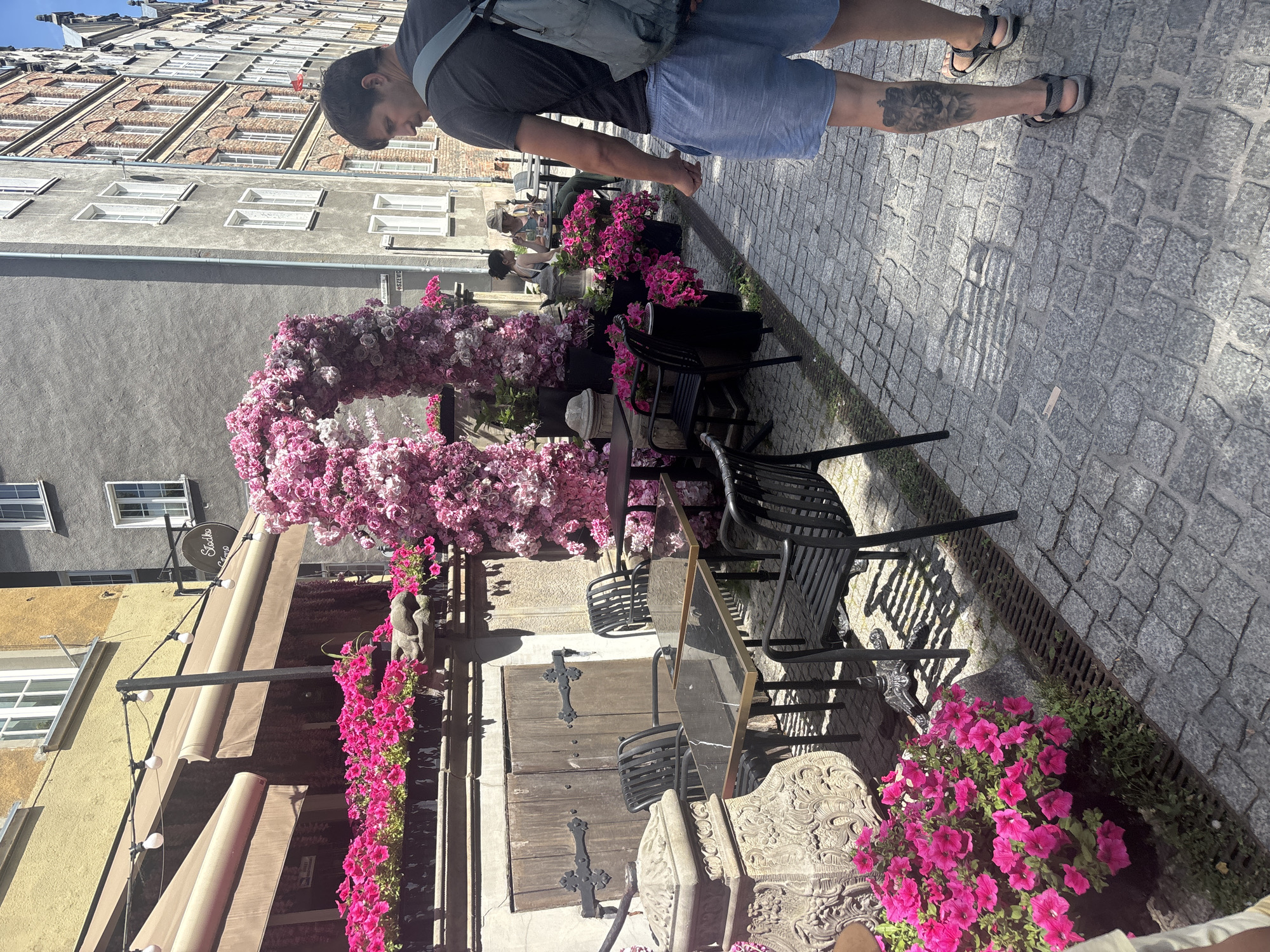 The image shows a street scene with a person standing near a building adorned with pink flowers. The person, wearing a dark t-shirt, blue shorts, and sandals, is partially visible from the waist down and is looking downwards. They have a backpack slung over their shoulder and a tattoo on their left leg.
The setting appears to be an outdoor seating area for a cafe or restaurant. There are tables and chairs on a cobblestone street. A large floral archway, made of many small pink and white flowers, dominates the foreground and arches over the walkway. More pink flowers are planted in pots and beds around the seating area. The building in the background is old with a brick facade and many windows, suggesting a historic urban environment. There are awnings extending from the building over the outdoor area.
The time of day appears to be daytime, with bright sunlight casting shadows. The weather seems pleasant. A sign on the building reads "Studio Foto". The overall mood is bright and cheerful, possibly indicating a pleasant day for outdoor dining or visiting.