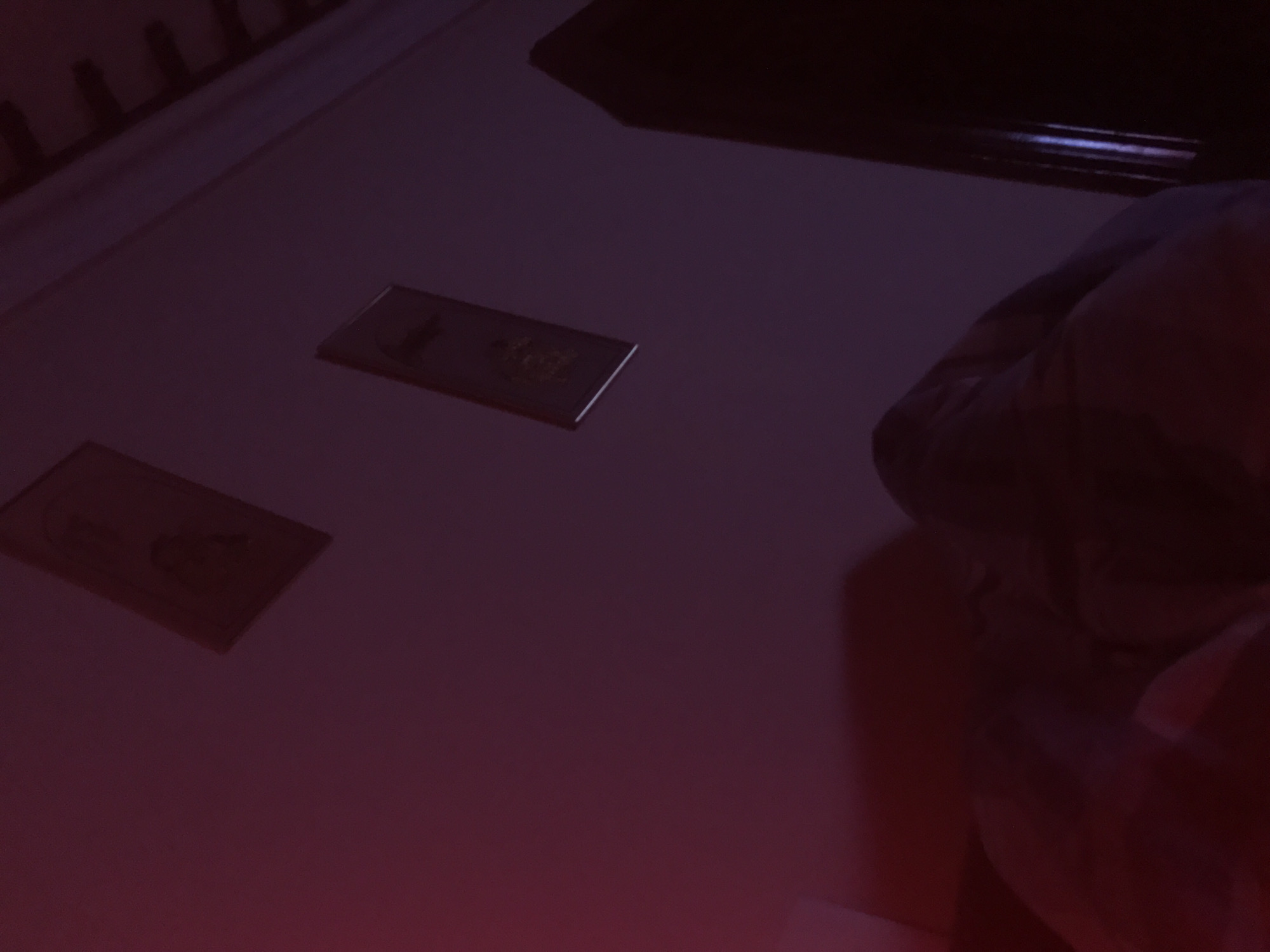 The image presents a low-angle view of a surface, likely a table or floor, illuminated with a dark red-purple hue. Two rectangular objects, possibly cards or small photographs, are positioned on the surface, with discernible images or designs on their faces. To the top right, there appears to be a partially visible human figure, seemingly shrouded in shadow or obscured from clear view. The background exhibits vague outlines of what appears to be furniture or architectural elements. There is no definitive information to suggest that the location is in Yerevan, Armenia.