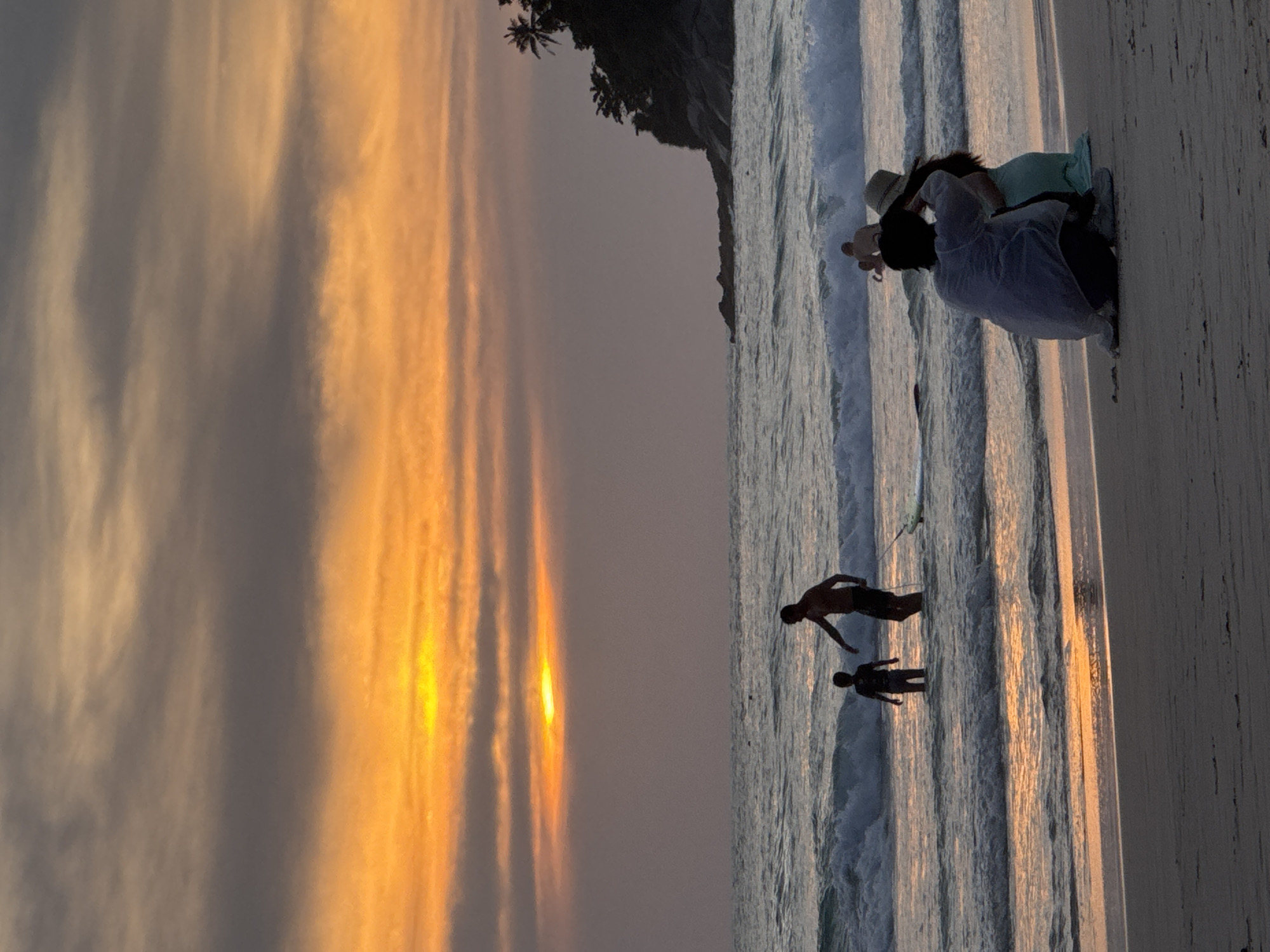The image depicts a sunset scene at Karon Beach, Thailand. The sky is filled with dramatic orange and gray clouds, with the sun partially visible through a break in the clouds, casting a warm glow on the water. Several people are visible on the beach and in the shallow water. In the foreground, a couple is seated on the sand, with one person taking a photo of the other. Further out in the water, a man and a child are standing, seemingly interacting. A surfboard is visible in the water near them. In the background, a rocky, tree-covered cliff juts out into the sea, and a few other figures are wading in the ocean. The waves are moderate, and the overall atmosphere is serene.