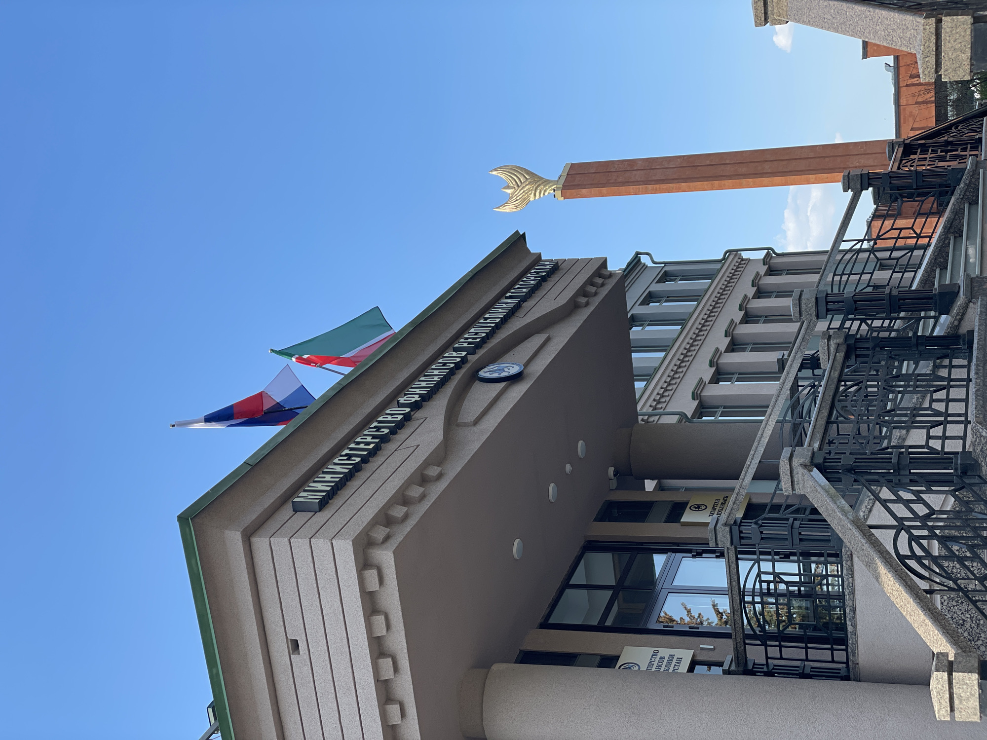 The image captures the Ministry of Finance of the Republic of Tatarstan. The building features the flags of Russia and Tatarstan. The monument of Zilant is visible in the background.