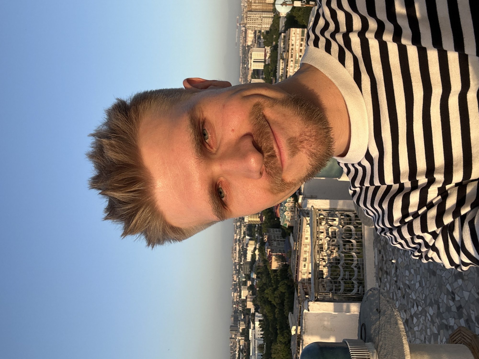 A man with blond hair and a short beard is on a rooftop in Tashkent, Uzbekistan. He is wearing a black and white striped shirt. The backdrop consists of buildings and a clear sky.