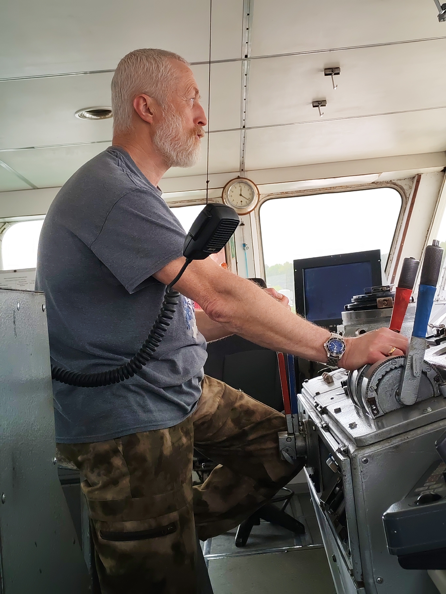 A man with gray hair and a beard is shown in profile, operating the controls of a ship. He wears a gray t-shirt and camouflage pants. The ship's controls feature levers with red and blue handles, and a gray metal housing. A clock is mounted on the wall behind him. A microphone is positioned near his face. The scene appears to be set within the ship's bridge. There is no information about the city of Gorodishche, Russia in this image.
