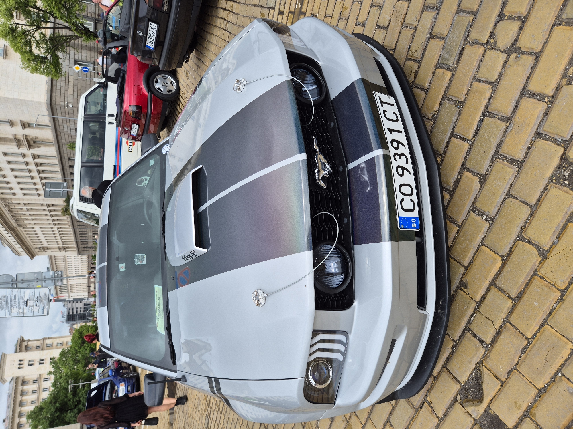 The image depicts a silver and black Ford Mustang with a Bulgarian license plate "CO 9391 CT" parked on a yellow brick-paved street. The street is in Sofia, Bulgaria, as suggested by the license plate. Other vehicles, including a red convertible and a white van, are visible in the background. People are seen near the Mustang and in the background. The surrounding environment includes buildings and trees, indicating an urban setting.