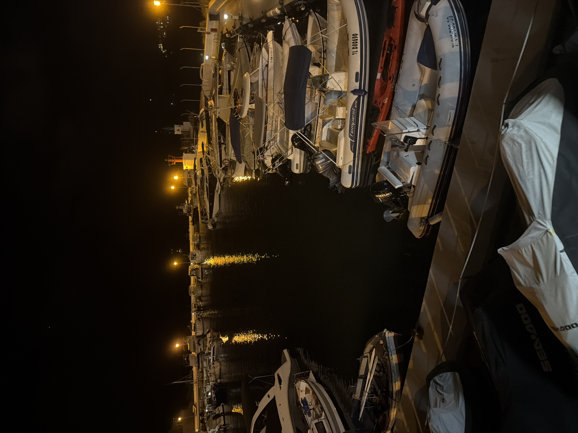 At night, in the city of Palmanova, Spain, a collection of inflatable boats and smaller vessels are moored in a harbor. The scene is illuminated by artificial lighting, casting reflections on the dark water. Several boats are partially covered with tarpaulins. In the foreground, a black Sea-Doo jet ski cover is visible. The overall environment appears to be a well-maintained marina.