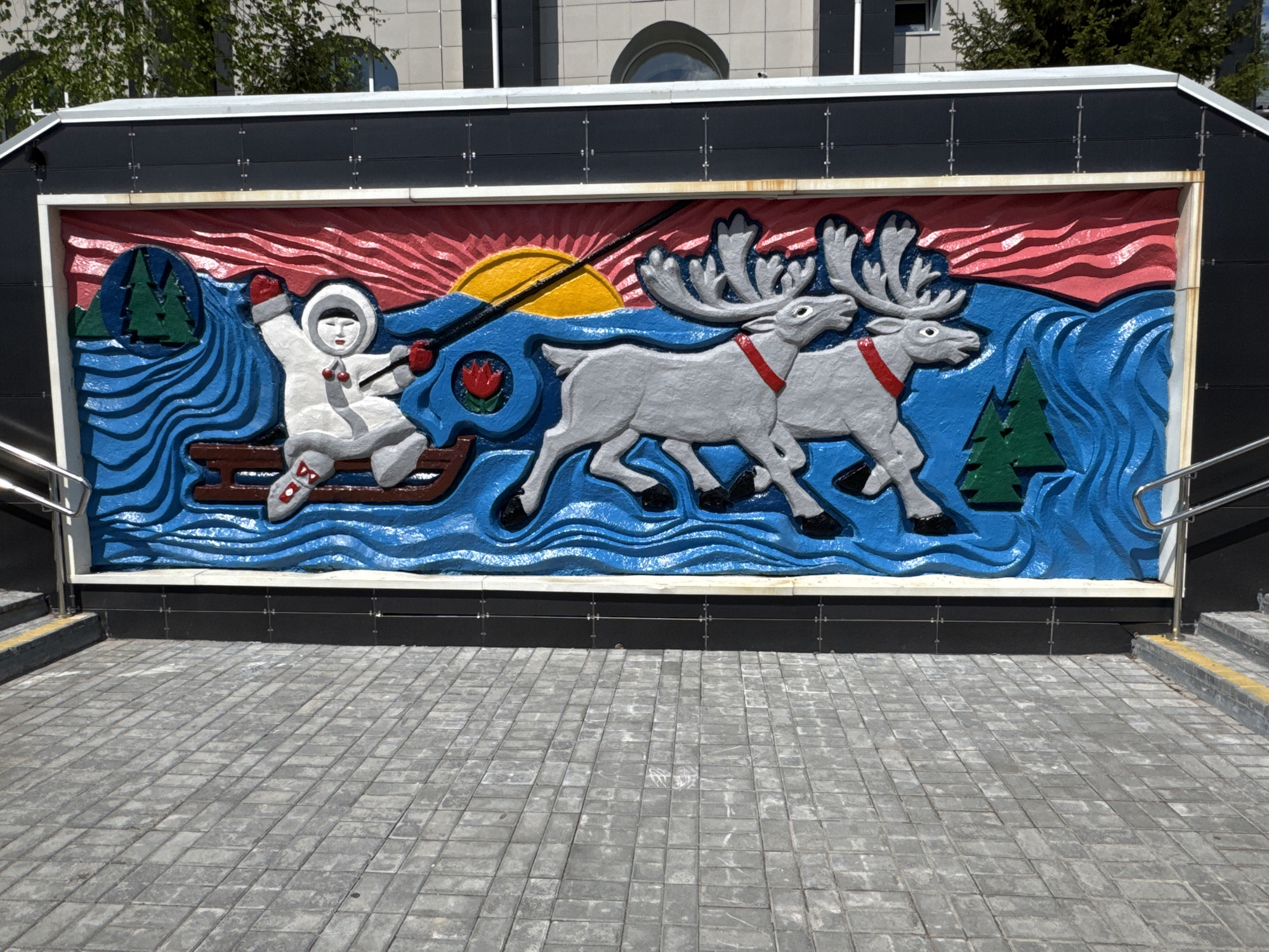 The image shows a bas-relief mural on the exterior of an underpass in Neryungri, Russia. The mural depicts an indigenous person in a white parka and fur-lined hood, seated in a wooden sled being pulled by two reindeer across a body of water. The scene is framed by a red sky with a yellow sun and is set against a backdrop of blue waves, green trees on the left, and a single green tree on the right. The person in the sled is holding reins and appears to be guiding the reindeer. The reindeer are grey with large antlers and red collars. The artwork is colorful and appears to be made of painted concrete or plaster. In the foreground, there is a paved walkway made of grey rectangular stones. Stairs and a metal handrail are visible on the right side of the image. The overall scene conveys a theme of Arctic life and travel.