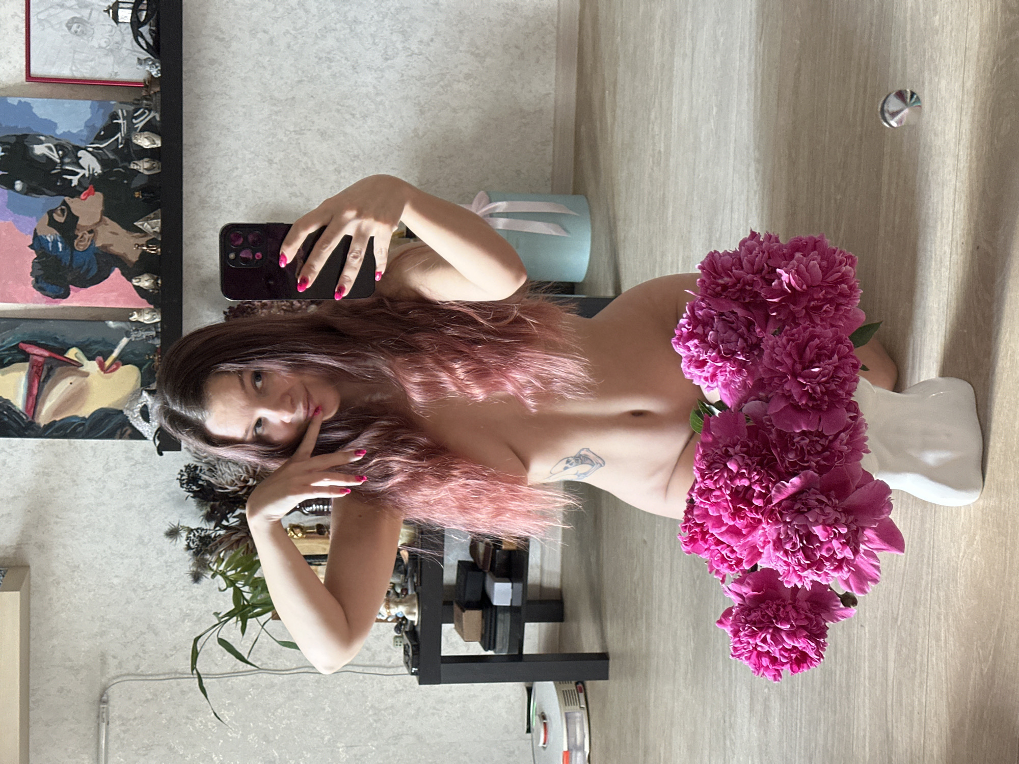 In a room with light-colored, textured walls and a light-wood floor, a young woman with long, pink-tinted hair is kneeling on the floor, holding a smartphone to take a selfie. She is naked, with a small tattoo on her left side. She is looking at the camera and posing with her hands near her face. In front of her, on the floor, is a white vase filled with a bouquet of bright pink peonies. To her left, a black side table is visible with various items on it, including a robotic vacuum cleaner on the floor beside it. In the background, a shelf displays several small figurines and artworks, including a painting of a woman smoking and a painting of Batman and Catwoman. To the right of the shelf, a light blue cylindrical object with white ribbons is present. The overall setting appears to be an indoor domestic environment.