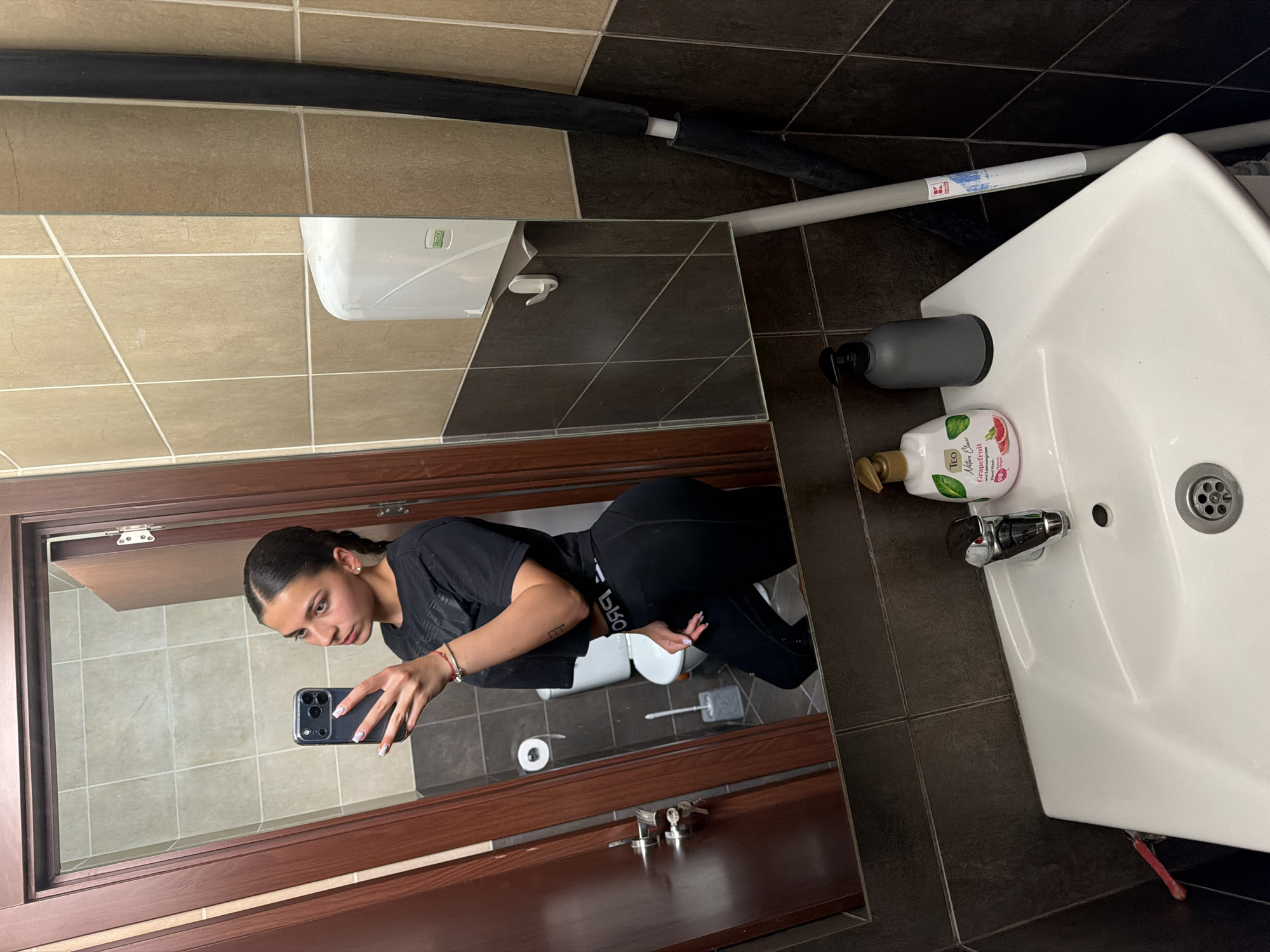 The image displays a mirror selfie taken in a tiled bathroom, likely a public or commercial restroom in Gabrovo, Bulgaria, with the entire photograph rotated 90 degrees clockwise.
In the mirror reflection, a young woman is visible from the waist up. She has dark hair styled in a sleek ponytail or braid and wears a black t-shirt and black leggings. She is holding a black smartphone with multiple camera lenses to take the selfie. Her fingernails are painted light with white tips, and she has a small, simple tattoo on her left forearm. Text on her leggings, partially visible, appears to be "бЬБО" (possibly Cyrillic). Behind her in the reflection, a white toilet, a toilet paper roll on a holder, and a toilet brush are visible.
The bathroom features a mix of light beige and dark brown square tiles on the walls, with dark brown tiles on the floor. In the foreground of the actual scene (outside the mirror), a white ceramic sink with a chrome faucet is prominent. Beside the sink are a grey pump soap dispenser and a bottle of "T.E.O Nature Olive Grapefruit Liquid Soap," which also states "Natural Origin," "98% Vegan," and "250 ml." A white "Liberty" brand hand dryer is mounted on the wall above the sink area. Various pipes, some insulated in black and others white or grey, run along the walls; one white pipe bears a red and blue logo. A wooden door frame surrounds the mirror. The woman's expression is neutral as she focuses on capturing the image.