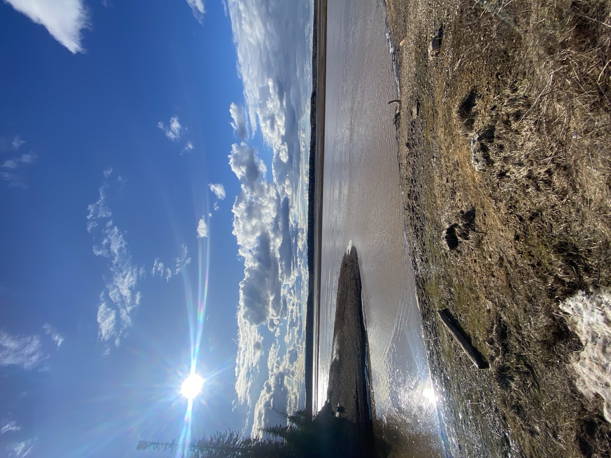 The image shows a landscape in Посёр, Russia. A wide river dominates the scene, its water reflecting the sunlight and clouds. A narrow strip of land or a sandbar extends into the river. On the right side of the river, there's a bank covered with vegetation. The sky is blue with scattered white clouds. The sun is shining brightly, creating a lens flare effect. The picture appears to be taken during the daytime.