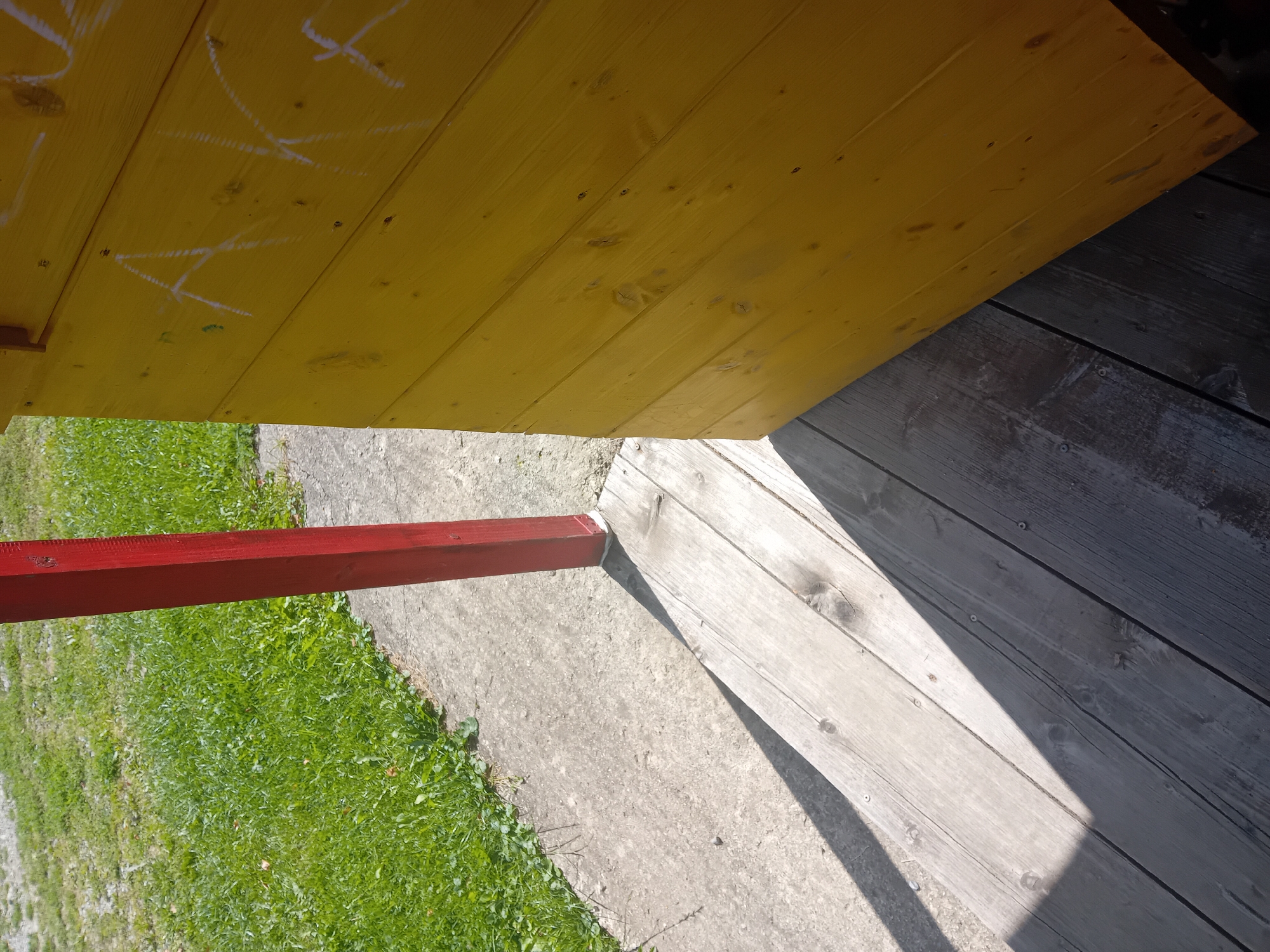 The image shows a close-up view of a wooden structure with yellow siding, partially obscured by a red post. White chalk or paint marks are visible on the yellow siding. The structure appears to be outdoors, with green grass and a concrete path in the background. A wooden walkway or deck is in the foreground, with a distinct shadow cast across it. The scene is likely part of a playground, park, or an outdoor recreational area in Zuberec, Slovakia. No people or animals are visible in the image.