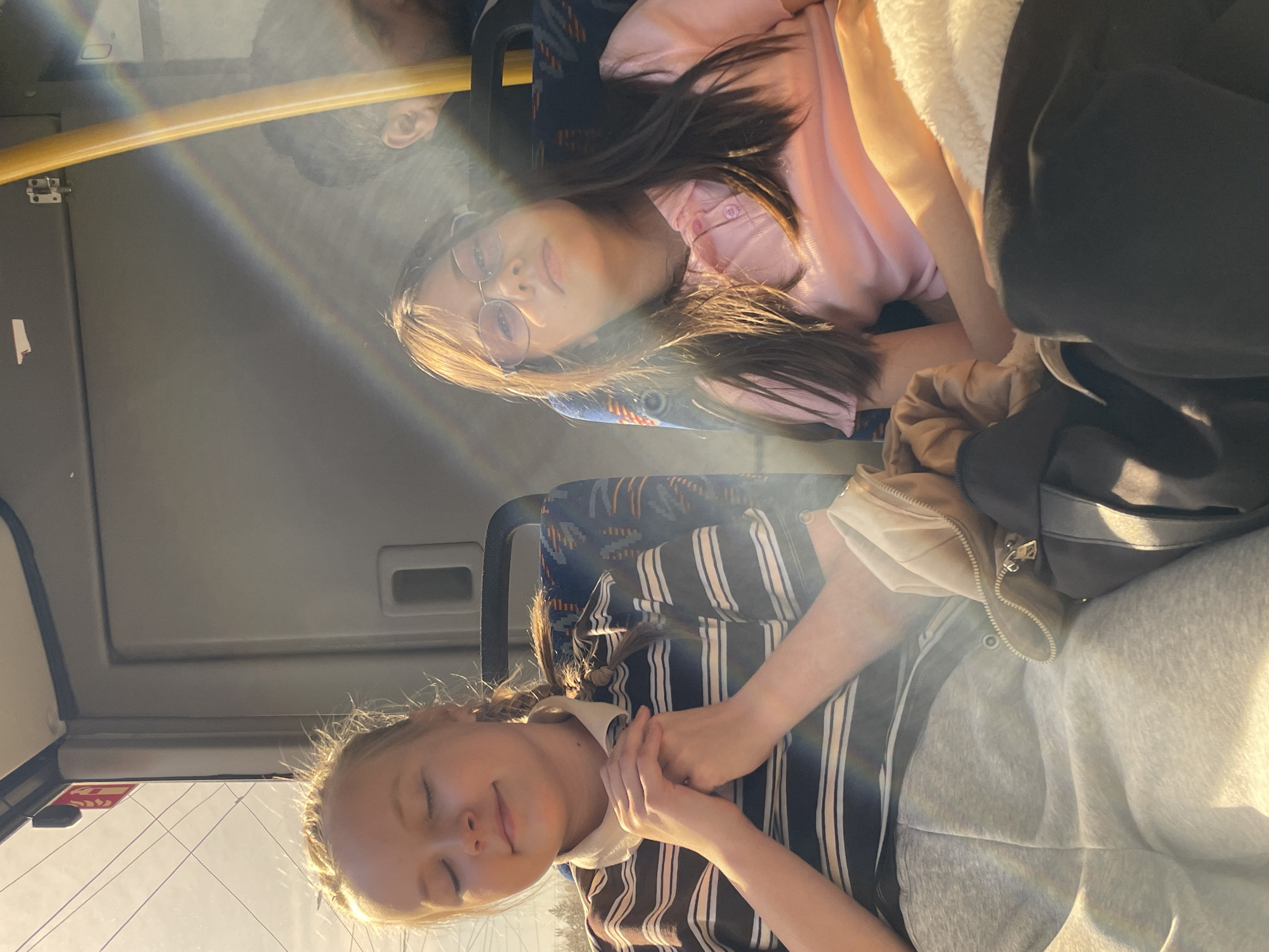 Two young women are riding a bus. The woman on the left has blonde hair pulled back in braids, is wearing a striped shirt with a white collar, and has her eyes closed. The woman on the right has long dark hair and is wearing glasses and a pink shirt. A yellow pole is visible near the top of the image. Light is streaming into the bus. The location appears to be inside a bus in a city, potentially Izhevsk, Russia.
