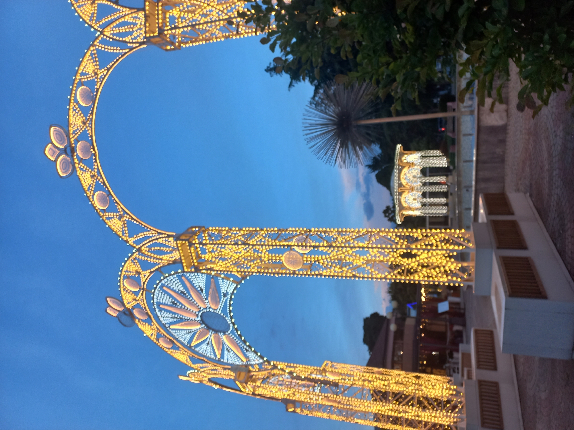 The image depicts an outdoor scene in Sochi, Russia, featuring decorative lighting at dusk. Dominating the foreground are two large, ornate light structures forming arched gateways, adorned with numerous small, warm-colored lights arranged in intricate patterns. Within these structures are decorative circles, some featuring floral designs. In the background, a modern dandelion-shaped sculpture stands tall beside a decorative building also illuminated with festive lighting. The sky transitions from light blue to darker hues, suggesting evening. The environment includes paving stones and a low wall with rectangular openings. The scene appears to be a public area or park decorated for an event or celebration.