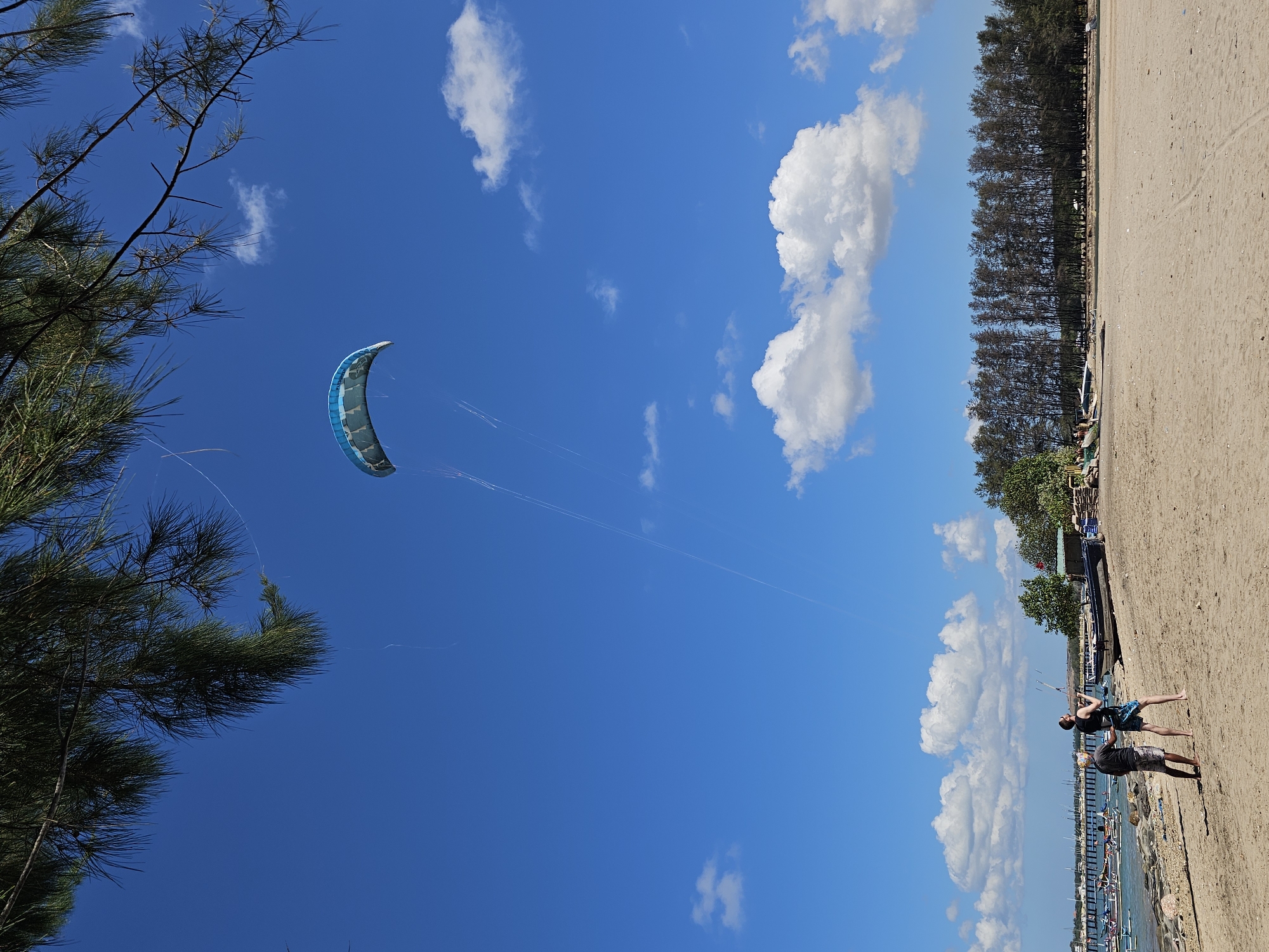 The image depicts a beach scene under a clear blue sky dotted with white clouds. In the foreground, two individuals are on the sand, with one appearing to be engaged in kite flying. A large blue and white kite is visible in the sky, controlled by lines held by the people. The beach is sandy, with some scattered rocks and debris. Further back, several boats are moored near the water's edge, and a pier extends into the sea. A dense line of trees forms the backdrop to the beach. This scene is likely located in Denpasar, Indonesia, a popular coastal city known for its beaches and water activities.