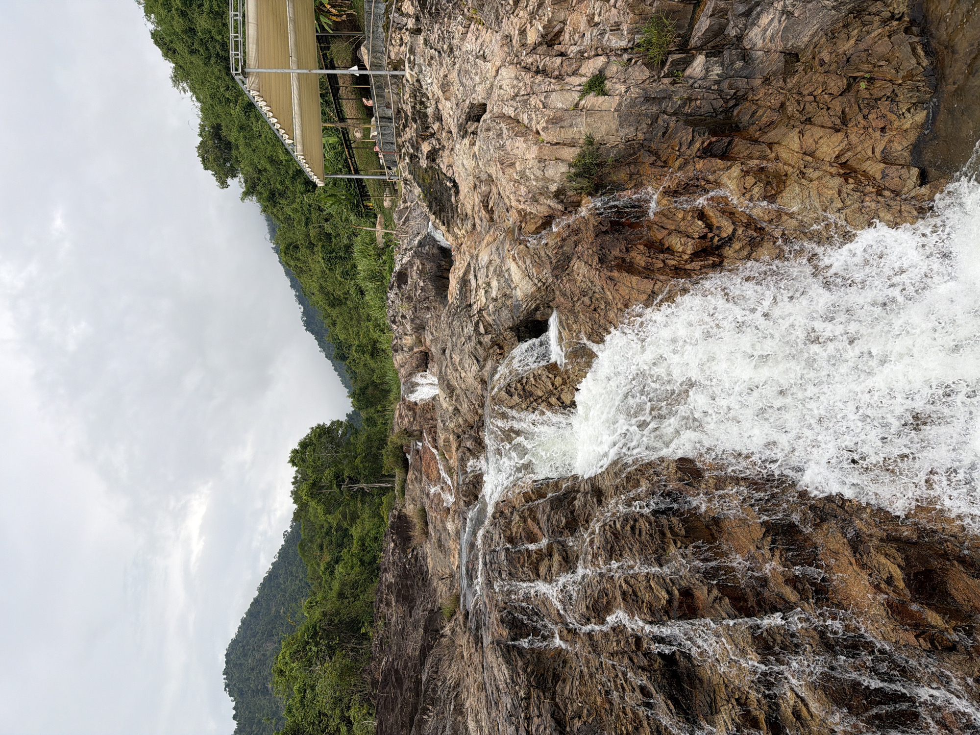 The image shows a waterfall cascading over brown and tan rocks in Khanh Vinh, Vietnam. Lush green vegetation covers a hill on the left side of the frame, contrasting with the rocky terrain. Above the waterfall, a structure with a tan canopy can be seen. The sky is overcast with grey clouds.