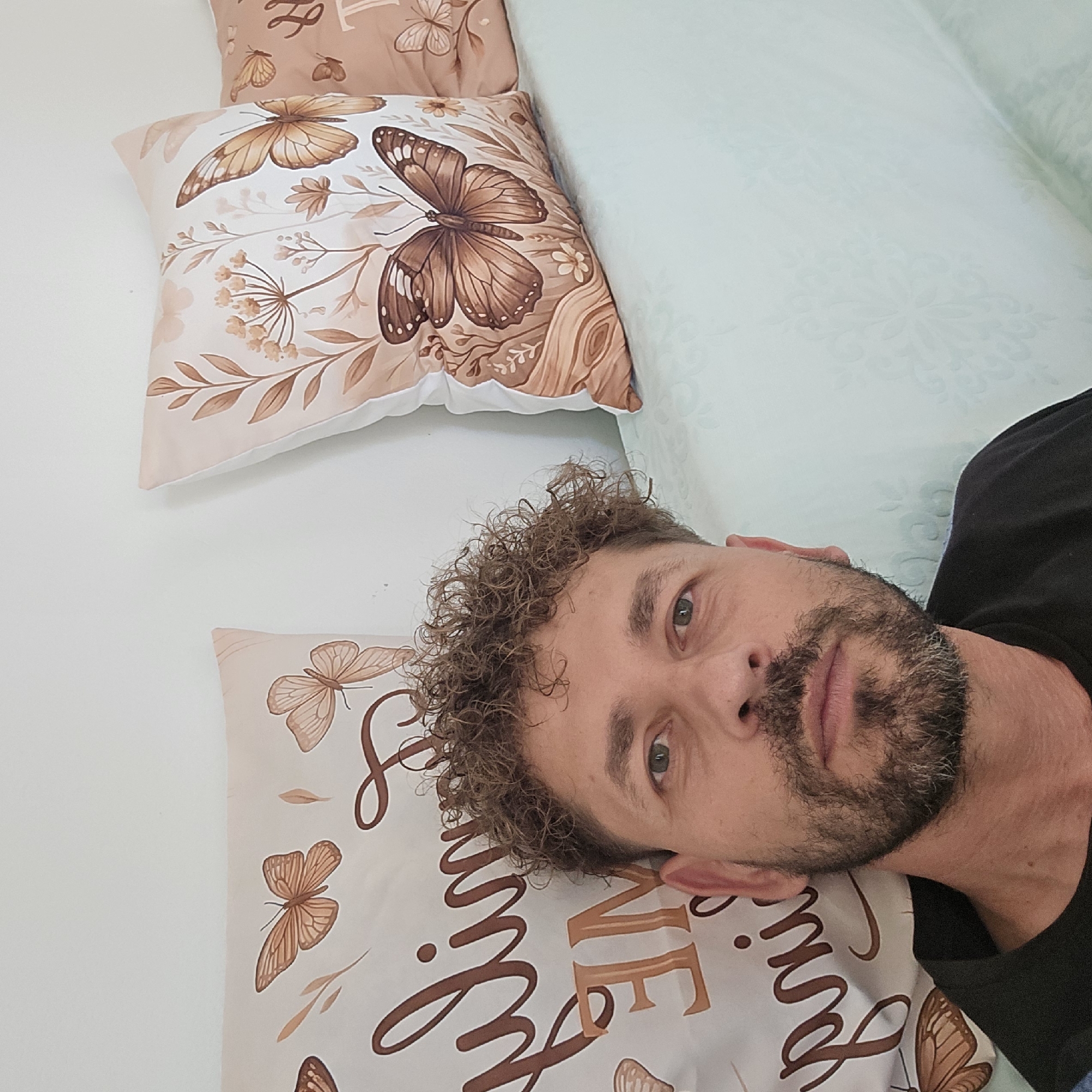 The image is a close-up, waist-up, eye-level selfie of a man with curly brown hair and a beard, taken indoors. He is looking directly at the camera. He is wearing a black t-shirt. Behind him, on the left, is a light-colored throw pillow with a brown butterfly and floral design, and text in brown script. To the right of that, and slightly further back, is another pillow with a similar brown butterfly and floral design. A light blue, patterned fabric, possibly a sofa or blanket, is visible in the foreground on the right, extending towards the man. The background is a plain white wall. The scene appears to be a domestic interior. There is no indication of the city of Athens, Greece, in the image itself.