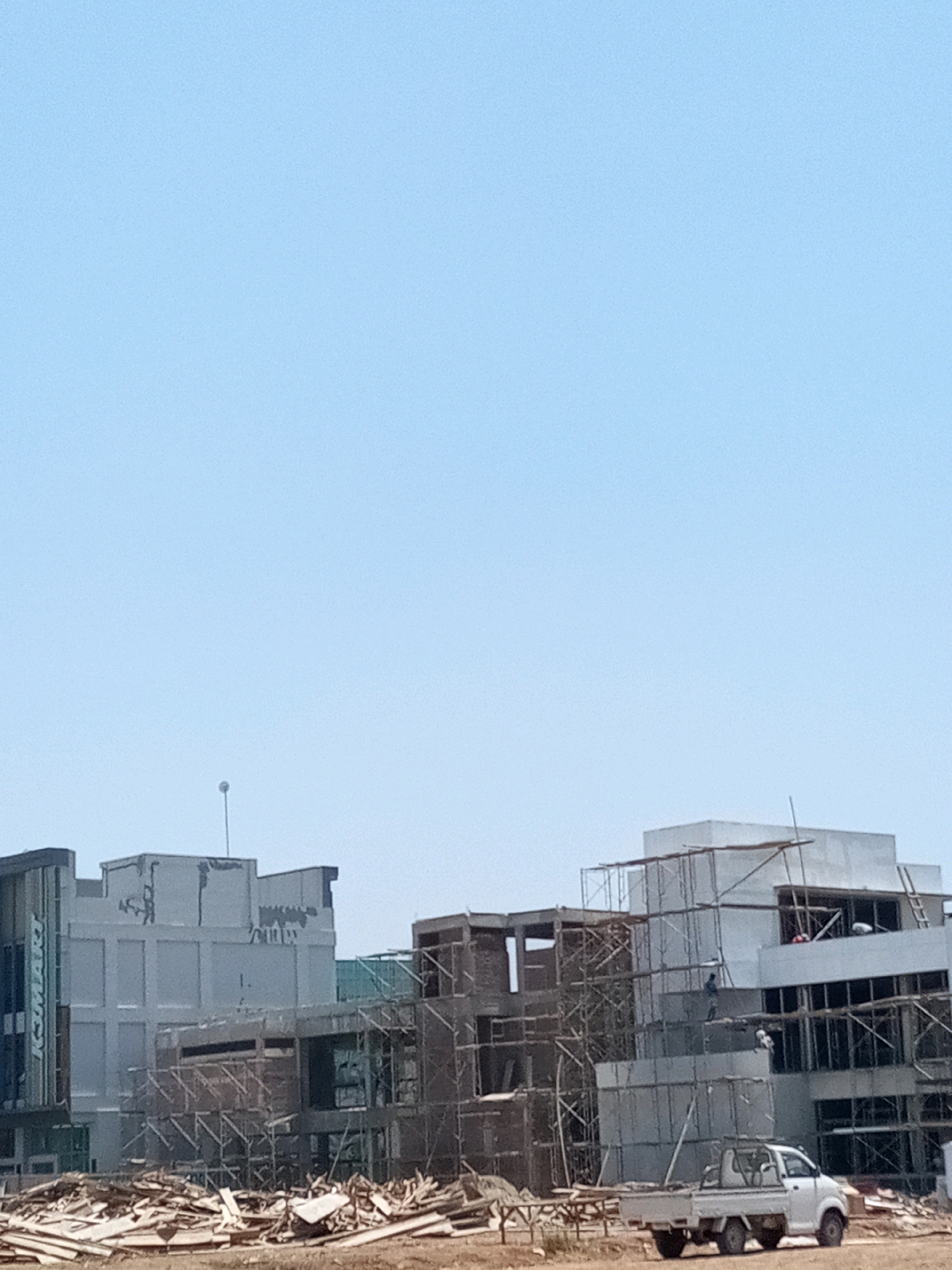 The image depicts an outdoor construction site in Bekasi, Indonesia, under a clear blue sky. In the foreground, a pile of wood debris sits on the ground. A white pickup truck is parked on a dirt road. Behind the debris and truck, a multi-story building is under construction. It features a modern architectural design with large windows and concrete elements. Scaffolding is erected around parts of the structure, indicating ongoing work. Several construction workers are visible on different levels of the building, engaged in their tasks. Some are standing, while others appear to be climbing or working at heights. Another building, appearing to be a completed section or a separate structure, is situated to the left of the construction site. This building has a sleek, contemporary facade with vertical signage that reads "KOMARI." The overall scene suggests active development and progress within a commercial or residential area.