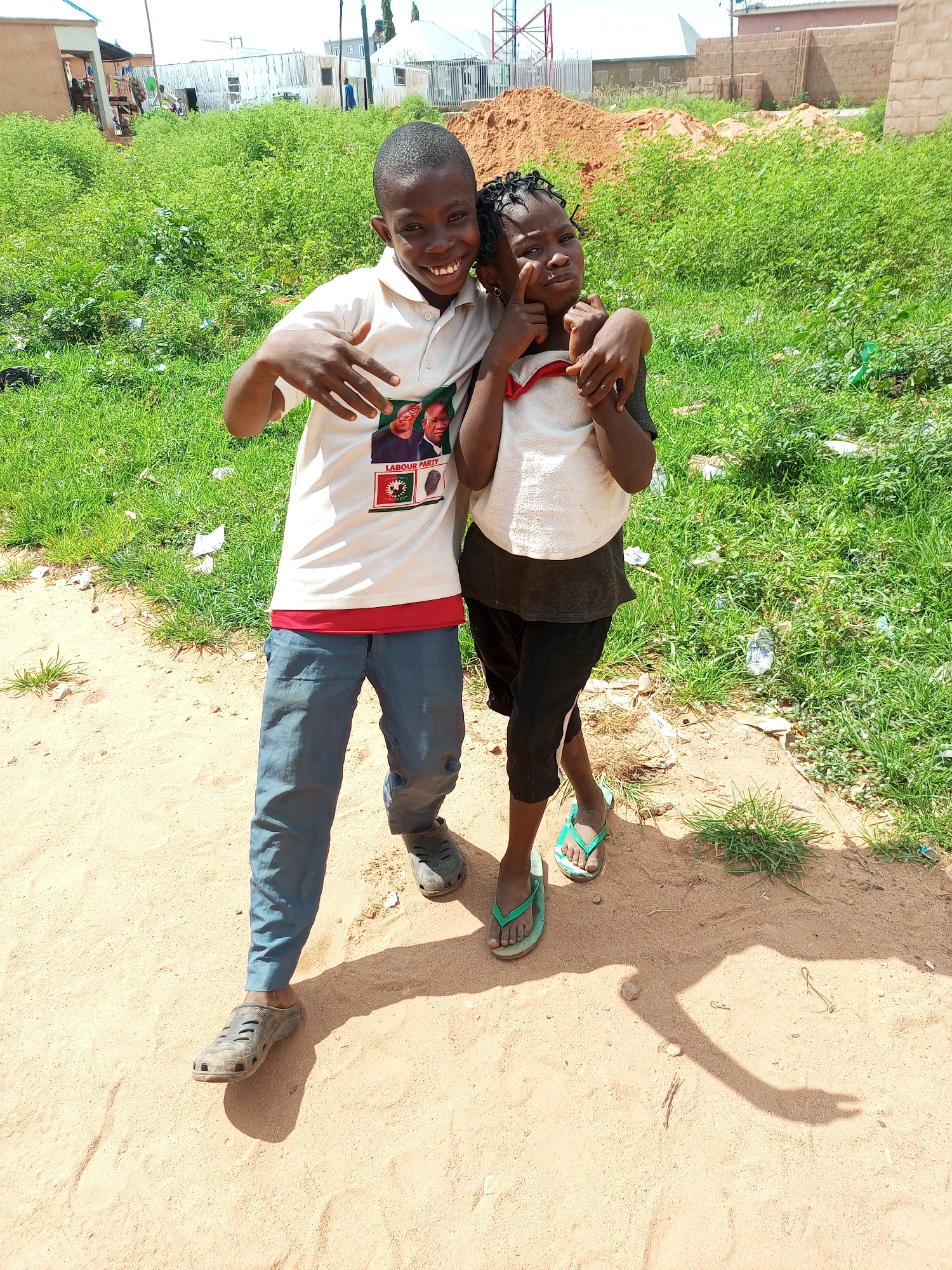 In Tempure, Nigeria, two children, a boy and a girl, pose for a photograph outdoors on a sandy, dirt ground with lush green vegetation and scattered debris in the background. The boy, on the left, wears a white polo shirt with a political campaign graphic featuring two men and the text "LABOUR PARTY", along with blue trousers and dark clogs. He smiles widely, gesturing with his hands towards the camera. The girl, on the right, has her arm around the boy's shoulders. She wears a dark t-shirt over a light-colored top and black shorts with a white stripe, paired with green flip-flops. Her expression is playful, with one hand near her cheek. Behind them, the environment suggests a semi-urban or developing area, with partially constructed buildings, mounds of reddish-brown earth, and a bright, sunny sky.