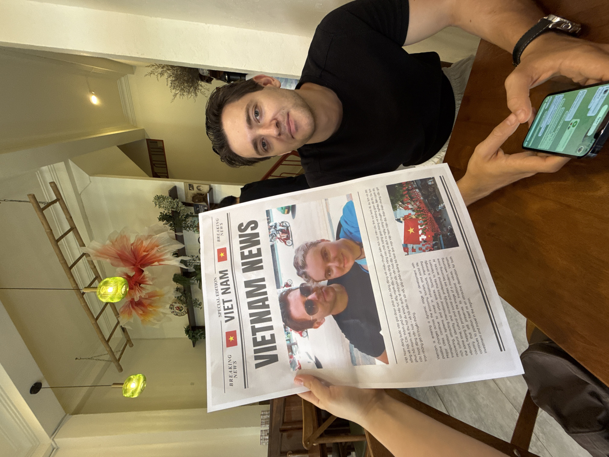 A man in a black t-shirt is seated at a wooden table, looking towards the camera with a calm, neutral expression. His right hand holds a smartphone, displaying a messaging application with green chat bubbles. In the foreground, another person's hand holds up a newspaper titled "VIETNAM NEWS," featuring "SPECIAL EDITION" and "VIET NAM" with a Vietnamese flag motif.
The newspaper includes a photograph of two men and articles in both Vietnamese and English. The text describes Vietnam's dynamic development, positive changes in its economy and tourism, infrastructure improvements, and its vibrant culture, portraying it as a "modern, welcoming country" and an "attractive destination."
The indoor setting features a wooden table and, in the background, various decorative elements. These include a large, elaborate floral wall art piece, two green pendant lights, a wooden ladder-like structure, and potted plants. The scene is located in Nha Trang Ward, Vietnam, which is consistent with the newspaper's content.
