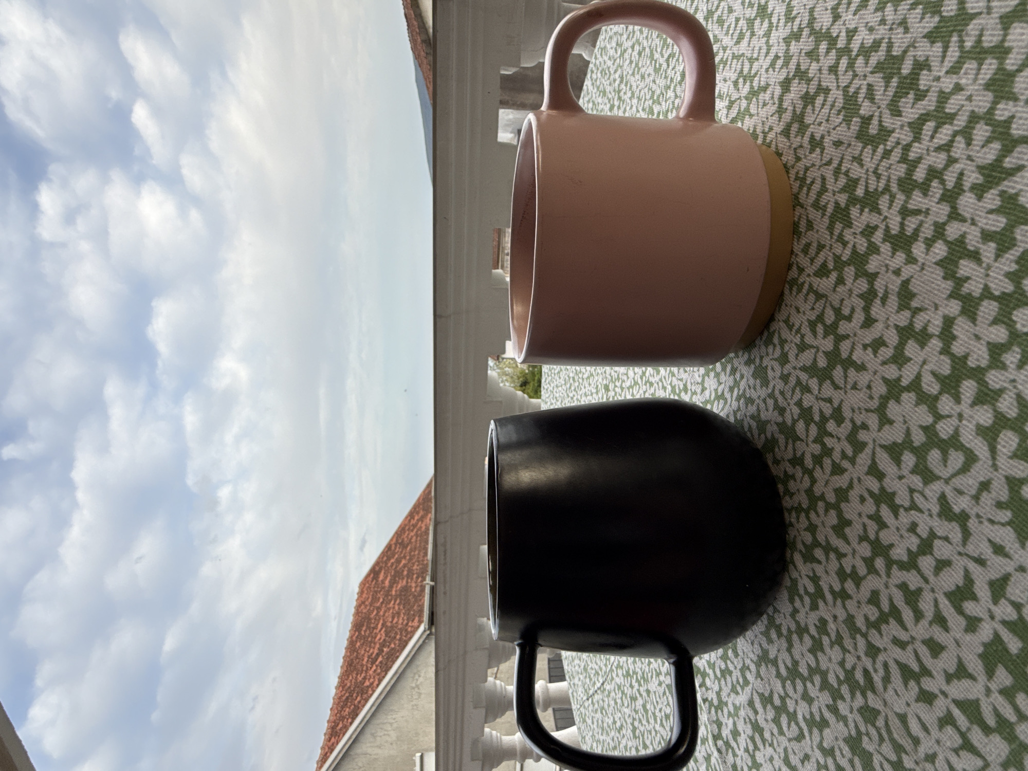 Two mugs, one pink and one black, are positioned side-by-side on a green floral patterned surface. Outside, there is a white ornate railing, a section of a house with a red tiled roof, and a view of a cloudy sky. The location is Bijela, Montenegro. No people are visible.