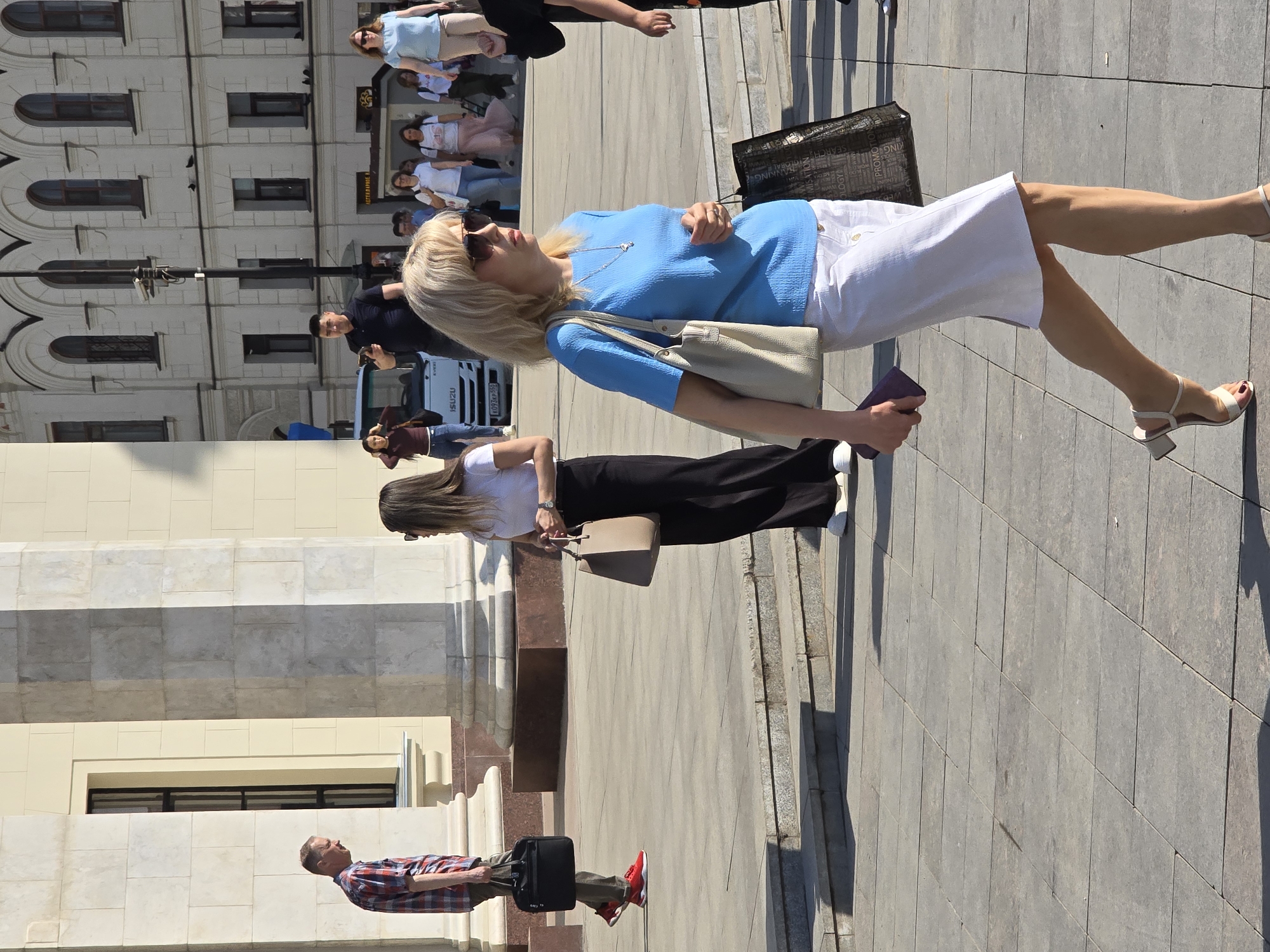 The image captures a daytime scene in Moscow, Russia. A light-skinned, blond-haired woman wearing sunglasses, a light blue short-sleeved shirt, and a white skirt is walking on a stone-paved sidewalk. She is carrying a beige tote bag on her shoulder and a small purple object in her hand. A slender, fair-skinned woman with long brown hair is walking in the same direction, wearing a white t-shirt and black pants, and carrying a beige shoulder bag. A man wearing a red and gray plaid shirt, khaki pants, and bright red shoes is walking in the opposite direction and pulling a black roller bag.
In the background, there is a light-colored building with arched windows and decorative elements. Further in the background, a group of people is standing near a white van. The overall environment appears to be an urban setting in Moscow.
