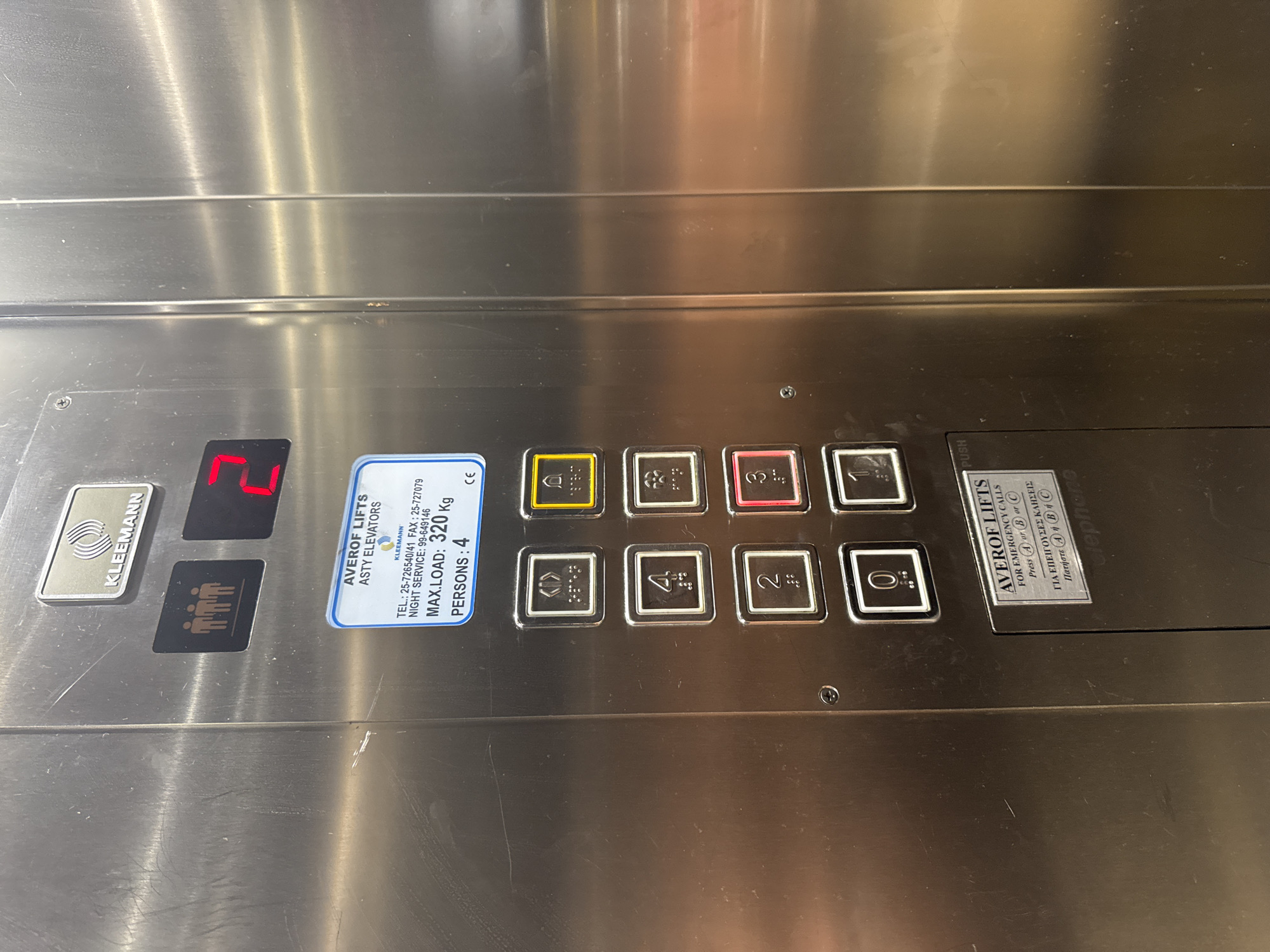 The image shows an elevator control panel inside a modern building. The panel is made of brushed stainless steel. At the top, there is a logo with the text "KLEEMANN". Below the logo, there is a black button with three stylized human figures, indicating a maximum capacity. To the right of this button is a red digital display showing the number "2".
Below the display, there is a white sticker with blue lettering that reads "AVEROFLIFTS ASTY ELEVATORS" and "KLEEMANN". It also provides contact information: "TEL.: 25-726540/41 FAX: 25-727079 NIGHT SERVICE: 99-649146". Further details on the sticker indicate "MAX.LOAD: 320 Kg" and "PERSONS: 4", along with a "CE" certification mark.
Below the sticker is a column of circular elevator buttons. From top to bottom, these buttons are:
- A button with a symbol of an open door and Braille.
- A yellow button with a bell symbol and Braille, likely for alarm or door open.
- A button with the number "4" and Braille.
- A button with the number "3" illuminated in red, and Braille.
- A button with the number "2" and Braille.
- A button with the number "1" and Braille.
- A button with the number "0" and Braille.
There is another smaller metal plaque at the bottom of the panel, also with "AVEROFLIFTS" printed on it, stating "FOR EMERGENCY CALLS Press A or B or C". Below this, in Greek, it says "ΓΙΑ ΕΠΕΙΓΟΥΣΕΣ ΚΛΗΣΕΙΣ Πατήστε Α ή Β ή C".
The environment appears to be the interior of an elevator cabin, indicated by the control panel and the metallic finish of the walls. The presence of the "ASTY ELEVATORS" text on the sticker suggests a connection to the Agios Tychona Community in Cyprus. The scene is clean and well-maintained, typical of a commercial or residential building. No people are visible in the frame.