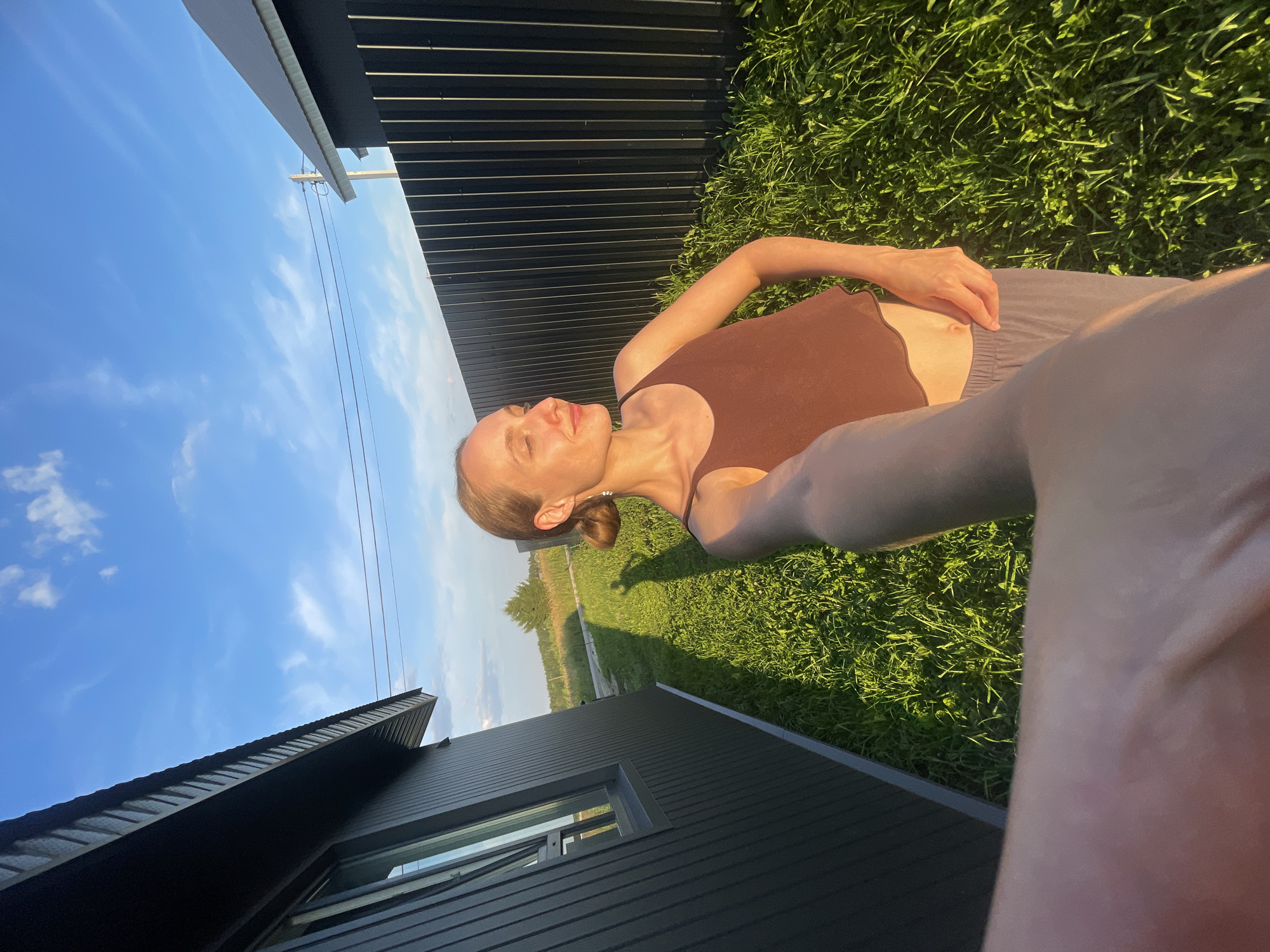 The image depicts a young woman taking a selfie outdoors on a sunny day, likely in a rural or suburban setting in the village of Podshivalovo, Russia. The woman is positioned in the foreground, facing slightly to her left, with her eyes closed and a gentle smile on her face. She is wearing a brown tank top and gray sweatpants. Her hair is pulled back in a bun.
Behind her, a dark gray, modern-style house with a sloped roof and a window is visible on the left. A tall, dark fence runs along the right side of the image. The ground is covered in lush green grass, with long shadows indicating the low angle of the sun. In the background, a power line pole and a distant tree can be seen under a bright blue sky with scattered white clouds. The overall impression is one of a relaxed, summer day.