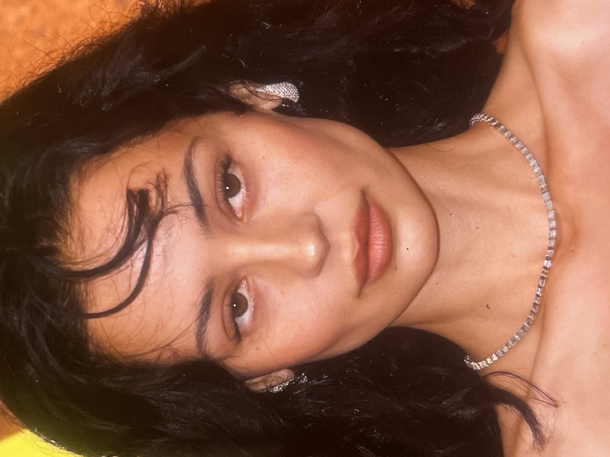 A close-up portrait shows a young woman, likely in Poltava, Ukraine, gazing directly at the viewer with a neutral to slightly alluring expression. Her head and upper torso are visible, positioned as if lying down or leaning back, with the image rotated ninety degrees counter-clockwise. She has dark, wavy hair cascading around her head and shoulders, with a few wisps framing her forehead. Her skin has a warm, natural tone, complemented by dark, expressive eyebrows and a hint of blush on her cheeks. Her brown eyes are open, and her lips are slightly parted, showing a natural lip color.
She wears two pieces of jewelry: a sparkling, possibly diamond, stud or cluster earring is visible in her left ear, nestled within her dark hair. A delicate, shimmering tennis necklace, appearing to be set with diamonds or rhinestones, adorns her neck. The background is softly blurred with warm, indistinct orange and yellow tones, suggesting an indoor setting. There is no discernible activity beyond posing for the photograph. No text is visible in the image.