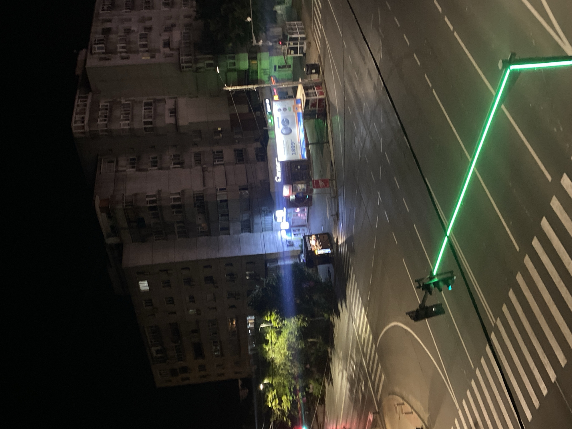 An aerial night-time view captures a city intersection in Iași, Romania. The scene is dominated by a large apartment building on the right, its windows illuminated with various lights. In the foreground, a crosswalk is visible with white stripes painted on the asphalt. A traffic light with a green signal is illuminated, casting a bright green glow across the road and a section of the crosswalk. To the left of the intersection, trees and foliage are visible, with streetlights illuminating the area. A kiosk or small shop is also present near the intersection, with some interior lighting. The overall atmosphere is urban and dimly lit, with the neon glow of the traffic signal being a prominent feature.
