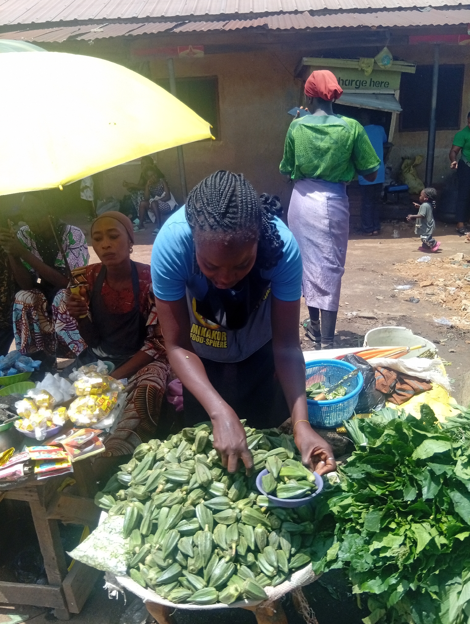 The image depicts a busy outdoor market scene in Bukuru, Nigeria. In the foreground, a woman wearing a blue t-shirt is sorting a large pile of okra. She is wearing a grey t-shirt with yellow text that reads "MINAKOR FOOD-SPHERE." To her left, another woman in a dark red top and a brown patterned apron is holding a knife and looking at the camera. In front of them are several small packages of yellow bouillon cubes, some plastic bags, and a blue basket containing some leafy greens. Behind the women, a woman in a green shirt and a purple skirt is visible, as is a child in a grey t-shirt and pink shoes. The background shows a simple structure with a corrugated metal roof, likely a shop or stall, with a sign that reads "charge here." The ground is dusty and appears to be unpaved. Several other individuals are present in the background, suggesting a lively market atmosphere. The lighting suggests it is daytime.