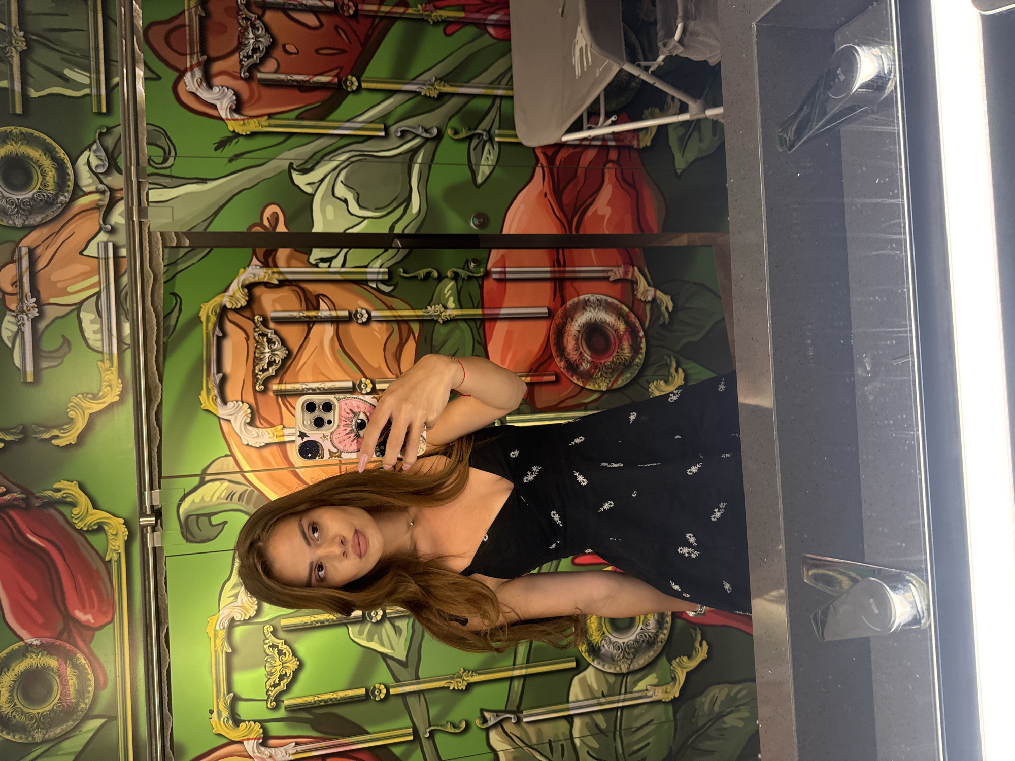 A woman with long, brown hair is taking a selfie in a bathroom with a vibrant mural on the wall. She is wearing a black dress with a floral pattern and is holding up her phone to take the picture. The mural features large, stylized flowers and ornate frames. A sink is visible in the foreground. The scene appears to be in a public restroom, possibly in a bar or restaurant. The location is identified as Plovdiv, Bulgaria.