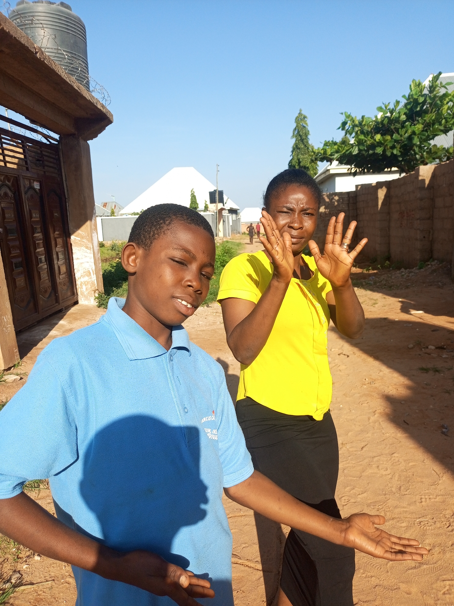 The image depicts two individuals in an outdoor setting, likely within a residential area in Tempure, Nigeria. A young boy, wearing a blue polo shirt, is positioned in the foreground on the left, facing slightly to the right with his eyes closed and mouth open in a smile. Behind him and to the right stands a woman wearing a bright yellow short-sleeved shirt and a black skirt. She is facing the camera with her hands raised, palms outward, in a gesture that could be interpreted as a greeting or a posed stance.
The environment appears to be a dirt road or pathway with sandy soil. To the left of the frame, a section of a building with a dark wooden door is visible, suggesting a house or compound entrance. Above this structure, a large black water tank is mounted on a concrete platform, typical of water storage solutions in many Nigerian communities. In the background, various structures, including a white-tiled roof and brick walls, are present. A few trees and green foliage add to the rural or semi-urban character of the location. The sky is clear and blue, indicating fair weather conditions. The lighting suggests it is daytime, with the sun casting shadows on the ground and the subjects.