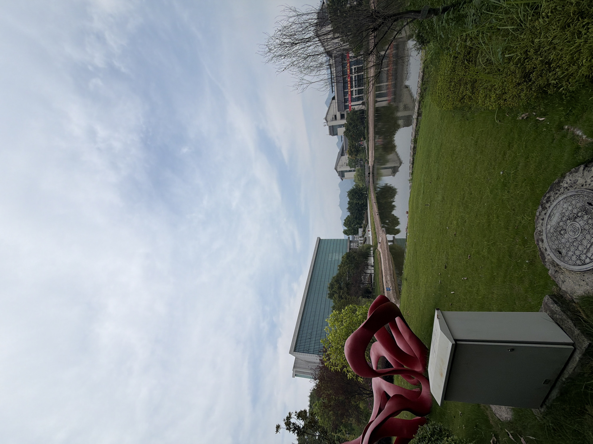 In Jiangjin, China, an outdoor scene unfolds with a large, modern building featuring a glass facade dominating the background. In the foreground, a vibrant red, abstract sculpture rests on a lush green lawn, with a gray utility box situated nearby. A calm body of water reflects the surrounding architecture and trees. The sky above is partially cloudy.