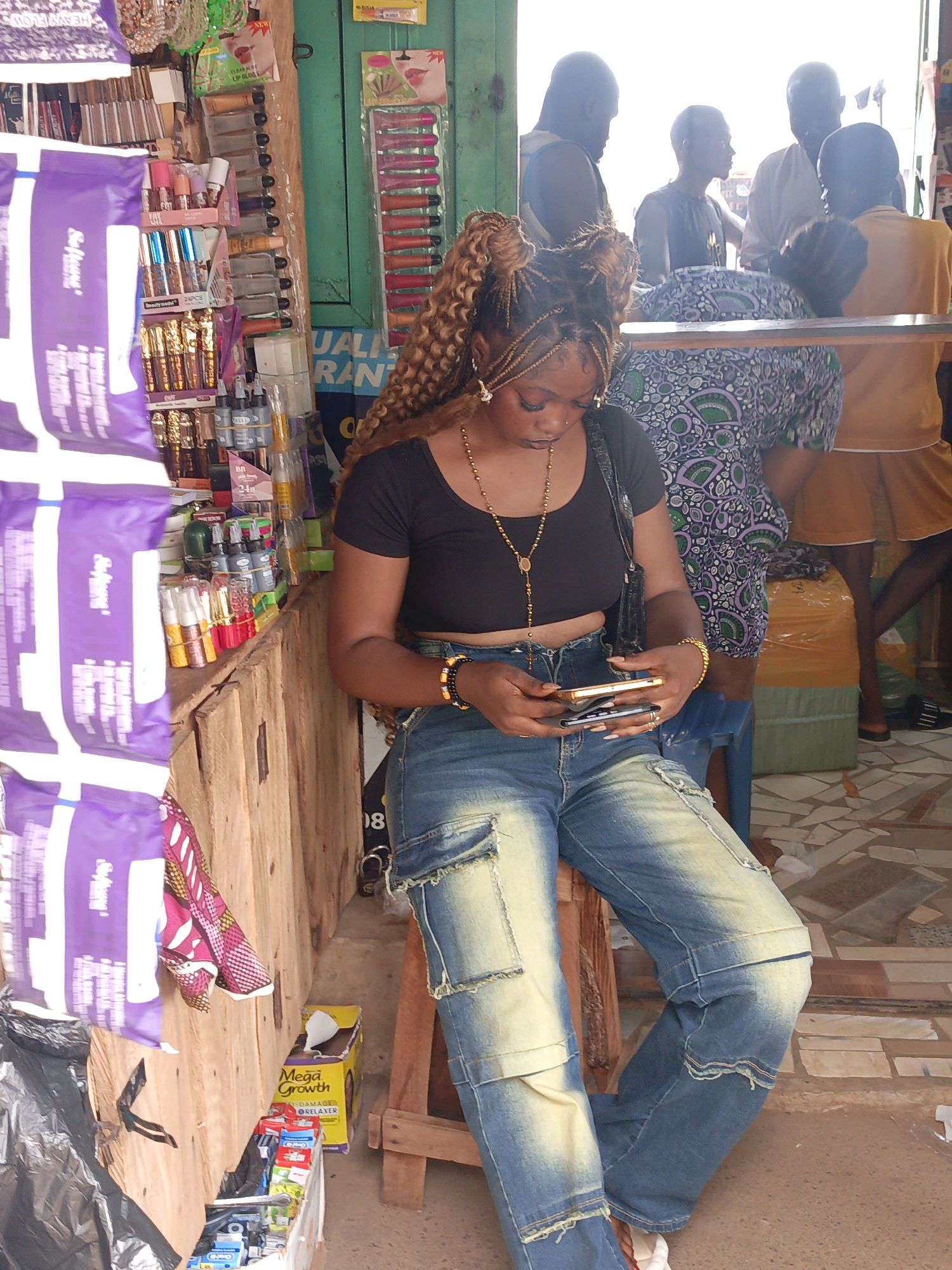 A young woman with braided hair sits on a wooden stool, absorbed in using two smartphones in what appears to be a retail store or market in Akwanga, Nigeria. She wears a black crop top, blue cargo jeans, and gold jewelry (necklace and bracelets).
The setting is a small retail space filled with various products: personal care items (lip glosses, hair relaxers), packaged goods, and cosmetic products. Some items are displayed on a wooden shelf, while others are hung on the wall. In the background, several other individuals stand near the entrance, indicating activity within the space. The floor has a patterned tile design, and light enters from an opening behind the people in the background. The store has a green painted trim and the items are priced.