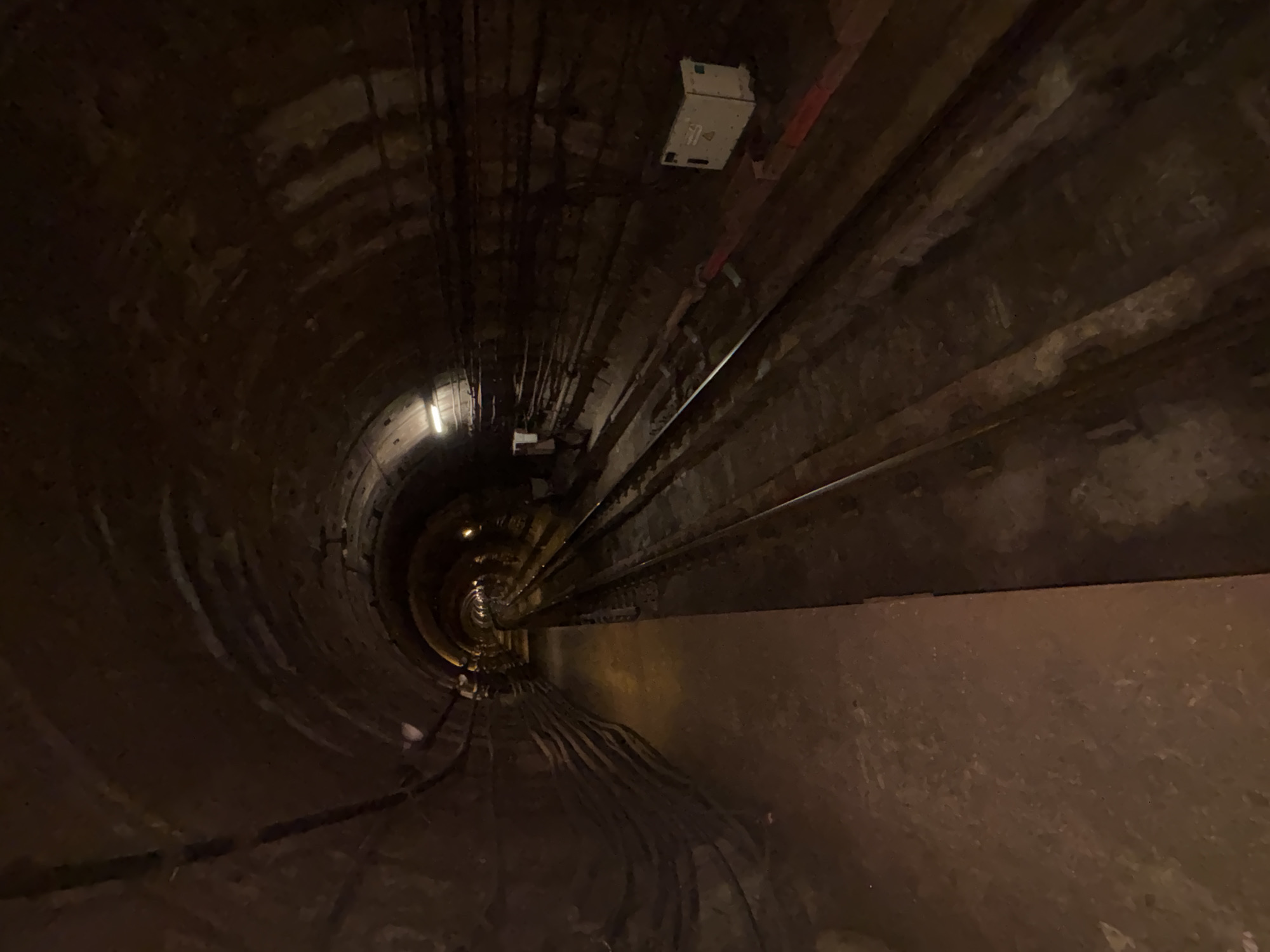 The image shows an empty, dark, arched tunnel, likely a subway or service tunnel, with railroad tracks running through it. The tunnel walls are constructed of rough, textured material, possibly concrete or brick, and appear worn and weathered, with some areas showing exposed pipes or conduits. Lighting is sparse, with a few visible light fixtures casting a dim glow. On the right side of the tunnel, a white electrical box is mounted on the wall. The perspective is from a low angle, looking down the length of the tunnel as it recedes into darkness. The overall impression is one of an industrial or utilitarian space, likely in an underground setting. Without further information, it's impossible to pinpoint this location to a specific place in Saint Petersburg, Russia, though its characteristics are consistent with infrastructure found in large cities.