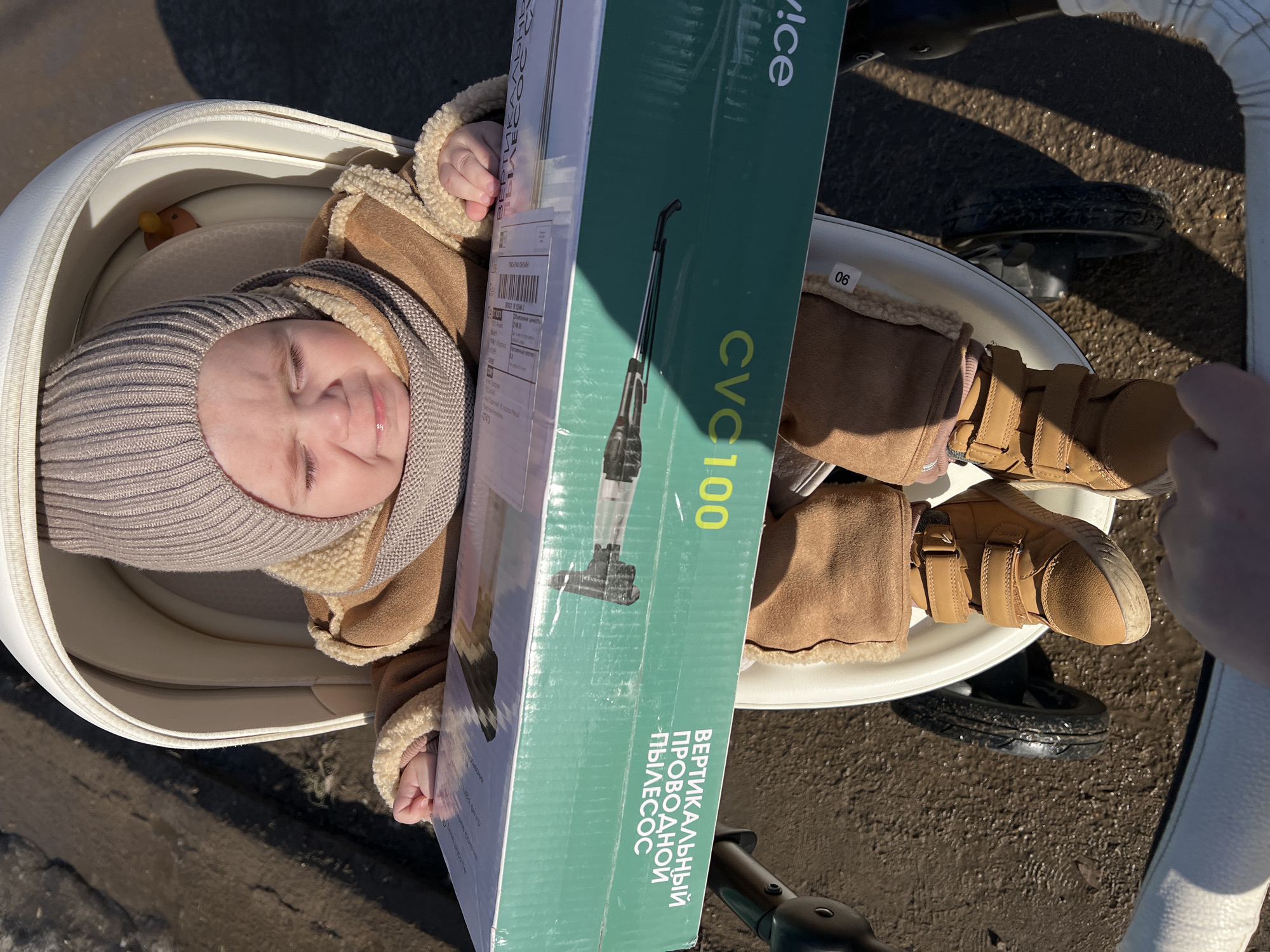 A baby wearing a beige jacket, hat, and shoes is lying in a white stroller in an outdoor setting. A box for a "CVC 100" vertical cordless vacuum cleaner is placed across the stroller. A label on the box reads "ПОЧТА РОССИИ" (Post of Russia) and indicates the city of Noviy, Russia as part of the address. The ground surrounding the stroller appears to be damp or wet. The image is taken from an overhead perspective.