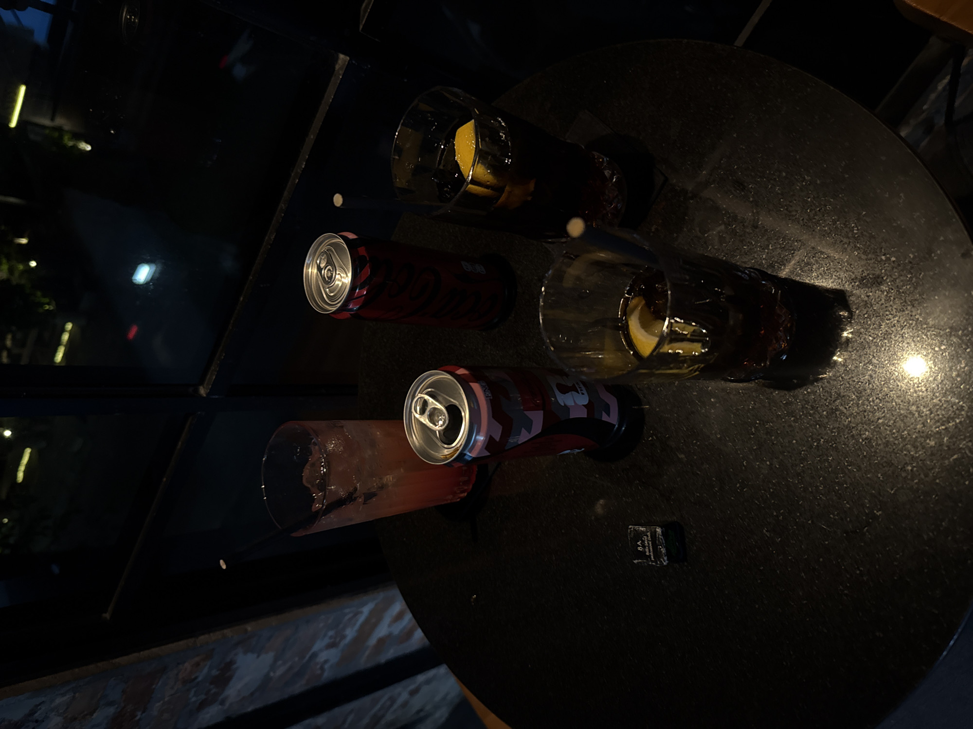 The rotated image captures a nocturnal scene, likely in a bar or restaurant. A dark, circular table with a subtle speckled texture, possibly granite, fills the lower and right portions of the frame, reflecting ambient light sources.
Arranged on the table are several drinks. Two tall glasses contain a dark beverage, each garnished with a lemon wedge and a thin white straw. Next to these are two opened soda cans. One can is clearly identifiable as Coca-Cola Zero Sugar, featuring a distinctive red and black camouflage pattern, with "ZERO SUGAR" and "Coca-Cola" visible on its side. The second can is also red and appears to be another Coca-Cola product. Further to the left, a clear glass holds a light reddish, translucent drink, also with a white straw.
The background is dark and out of focus, showing blurred reflections of various lights, suggesting large windows looking out into a dark exterior or a dimly lit interior space. A small, dark rectangular object, possibly a coaster or label, rests on the table. Partially legible text on it includes "8.A" and some smaller script, which appears to say "Professional Use Only." No people are visible, emphasizing the drinks and the ambient setting.