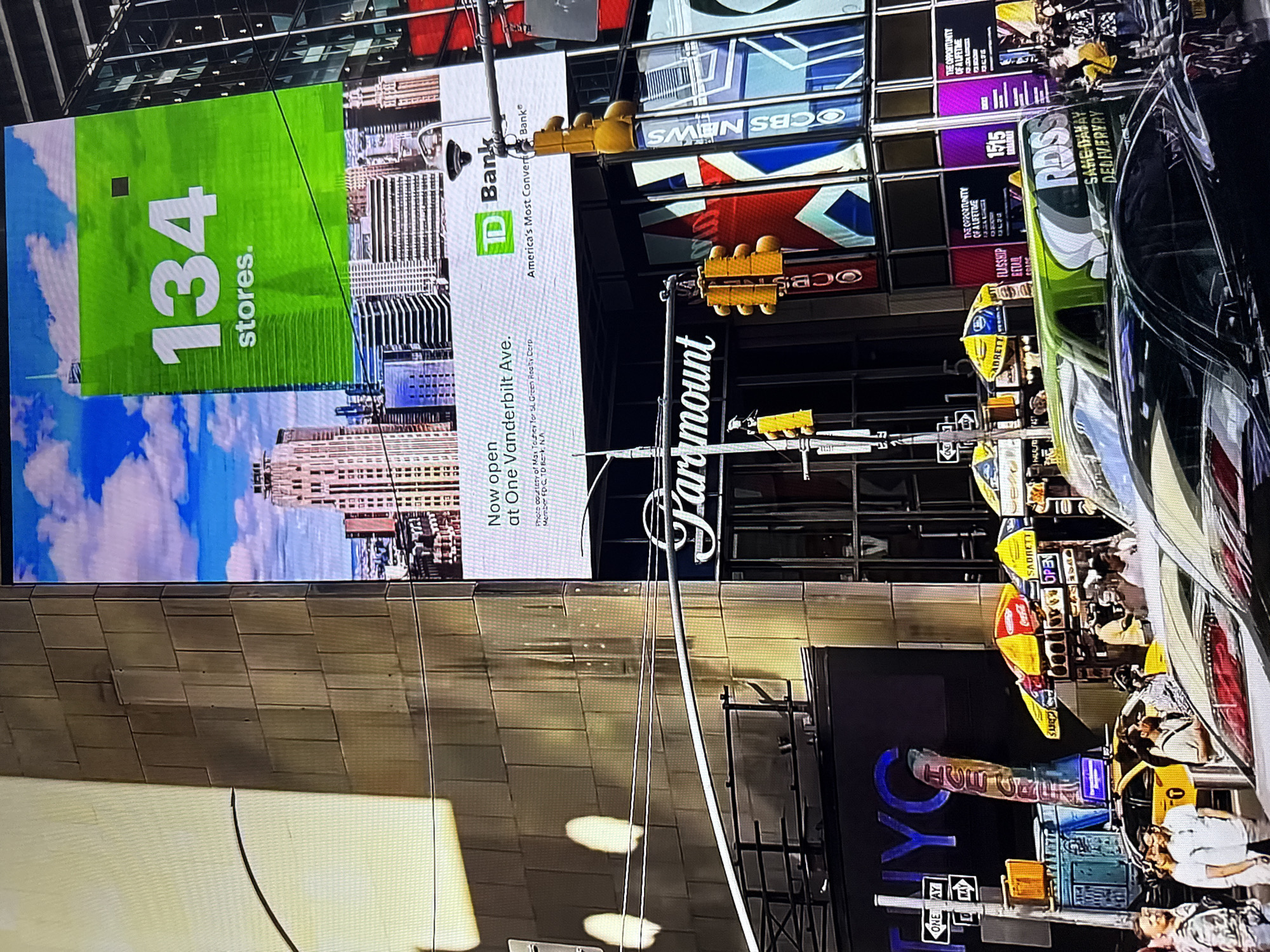 The image depicts a bustling urban scene, likely Times Square in New York City, based on prominent signage for "Paramount" and "CBS NEWS". A large digital billboard displays "134 stores." next to a TD Bank advertisement. Below, traffic lights and street signs are visible, along with yellow taxis and pedestrians. Food vendors with colorful umbrellas are also present, contributing to the lively atmosphere. The architecture suggests a dense city environment with tall buildings. The question asks to include the location in Burgas, Bulgaria, but the visual evidence does not support this claim; all indicators point to a major city in the United States.