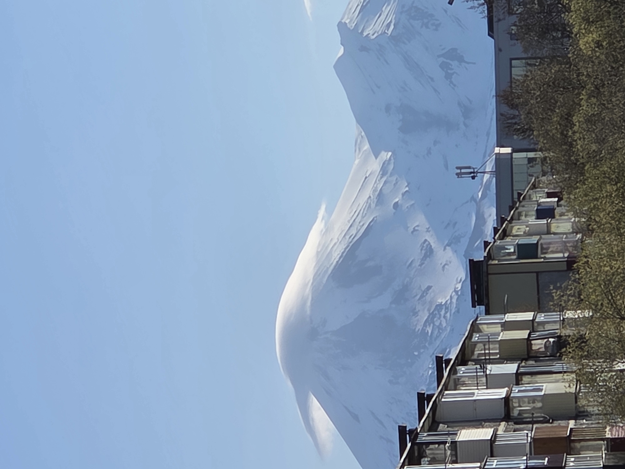 This image shows a mountain towering over buildings in the city of Unknown, Unknown. The mountain is covered in snow and has a large cloud formation extending down from the summit. In the foreground are densely packed apartment buildings with metal balconies. Trees and a building with a satellite dish antenna are visible to the right. The sky is a clear blue. The image appears to be rotated counterclockwise.
