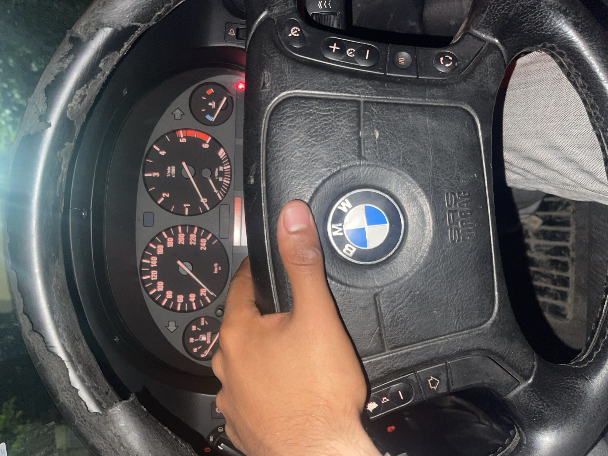 In Dolni Dabnik, Bulgaria, an individual's hand is gripping a damaged steering wheel of a car. The steering wheel exhibits significant wear and tear, with the leather covering peeling off in several places. The dashboard displays various gauges, including a speedometer, a tachometer, and a fuel gauge. The BMW logo is visible in the center of the steering wheel. The interior of the car appears to be dimly lit, suggesting it is either night or the car is in a shaded area. The overall condition of the steering wheel indicates age and extensive use.