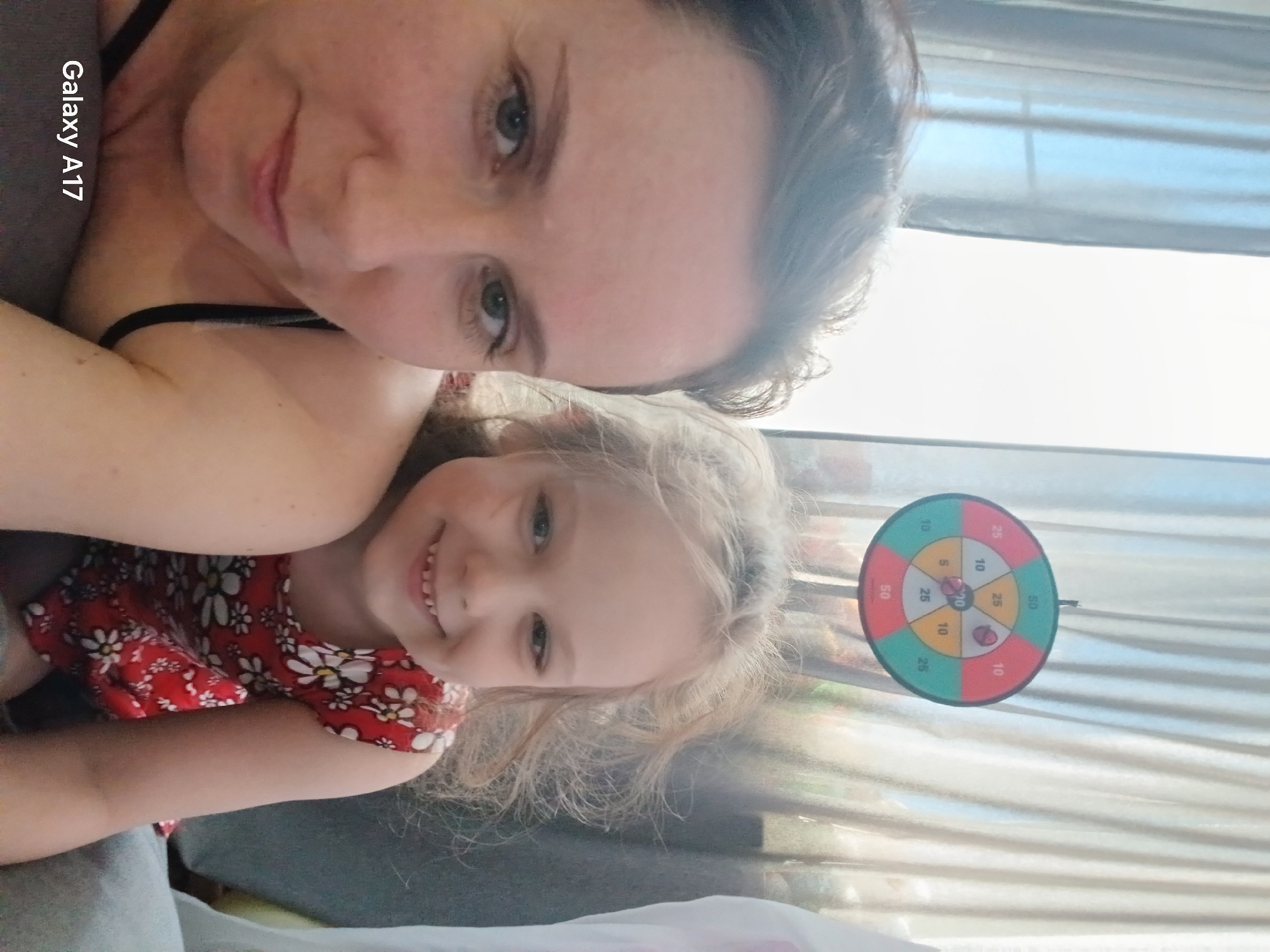 A selfie-style photo depicts an adult and a child, likely a mother and daughter. The adult has fair skin, light brown hair, and is wearing a dark-colored top with thin straps. The child, presumably a girl, has blonde hair and is wearing a red dress with white floral patterns. Both are smiling at the camera. A dartboard hangs on a wall with scores labeled. The background includes a window with sheer curtains. Text indicating "Galaxy A17" is visible. The location appears to be indoors, though there are no clues to specifically identify this as being in Yekaterinburg, Russia.