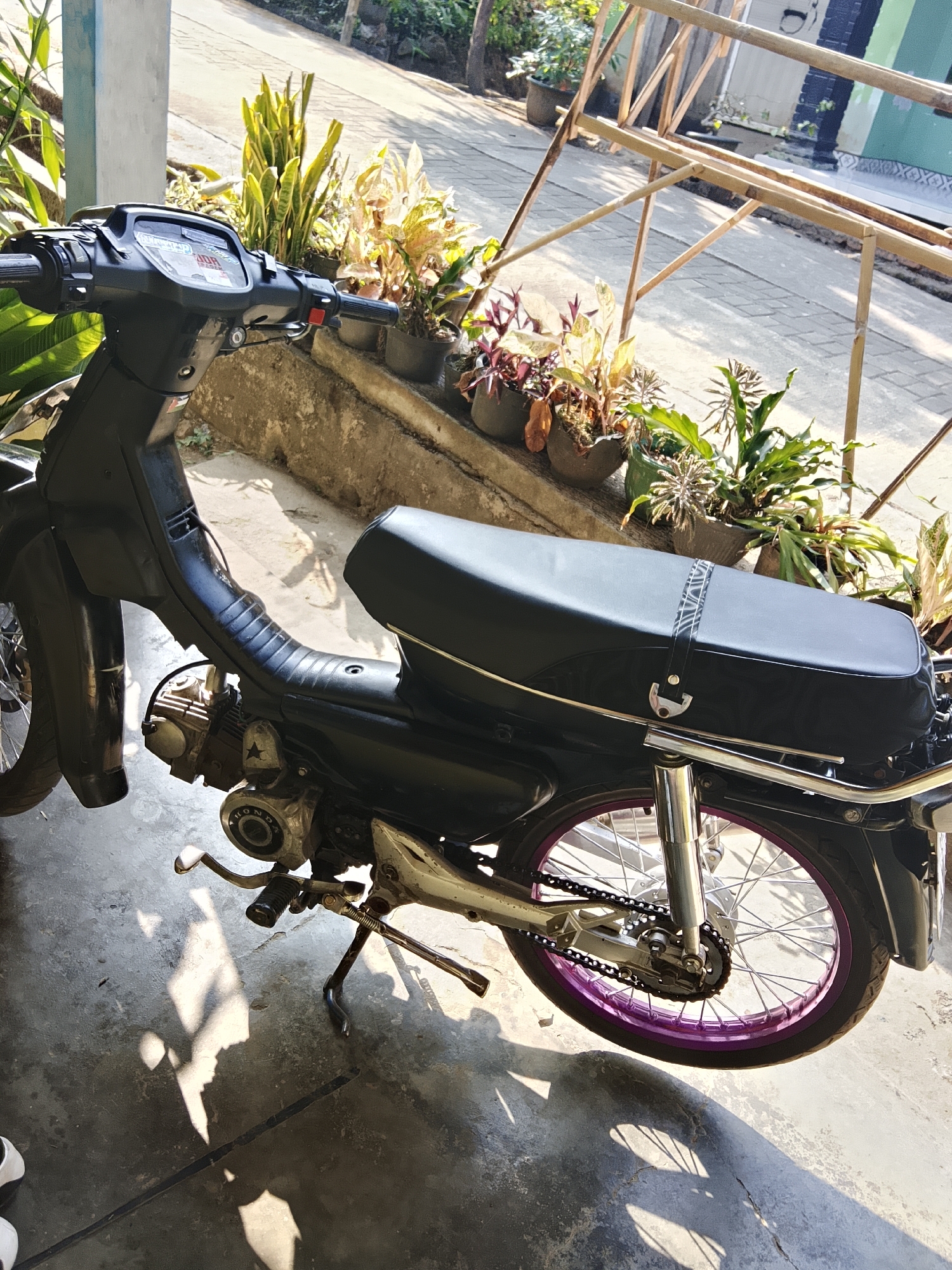 This is a medium-shot, eye-level, outdoor photograph of a black motorcycle parked on a concrete surface. The motorcycle is viewed from the right side, with its front partially obscured by a pillar. The motorcycle has a black seat with a decorative strap, a metallic gray engine, and a silver chain drive. The rear wheel has a purple rim. In the background, there are several potted plants with green and reddish foliage, placed on a concrete ledge. A bamboo structure is visible further back. Sunlight casts shadows on the ground and on the motorcycle. The scene appears to be a residential or semi-residential area, possibly a courtyard or alleyway, given the presence of the motorcycle, plants, and basic construction elements. The lighting suggests it is daytime, with bright sunlight. The location is identified as Tawangsari, Indonesia. No people are present in the frame.
