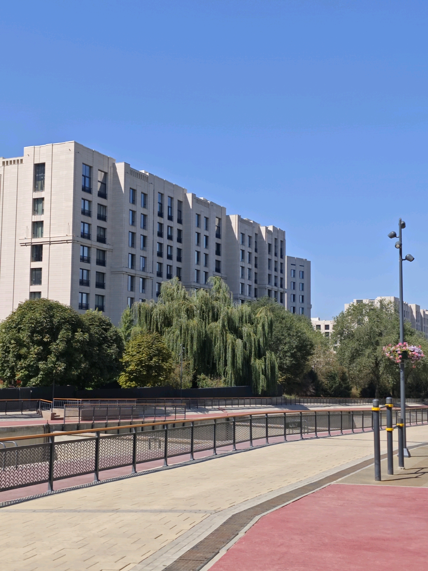 The image shows a modern, multi-story building with a light-colored facade, likely constructed from stone or similar material. The building features numerous windows with dark frames, arranged in a grid pattern across its expansive length. In front of the building, there are several mature trees, including a prominent weeping willow with its characteristic cascading branches, as well as other deciduous trees.
The foreground of the image depicts a well-maintained urban walkway or promenade. This area is paved with light-colored square tiles, with sections of red-colored material that could be a running track or designated pedestrian path. A dark metal fence with a decorative pattern separates the walkway from a lower level, which appears to be a canal or river. The fence has a wooden handrail along the top.
Further to the right, a tall, dark lamppost with multiple light fixtures is visible, and a hanging basket filled with pink and purple flowers is attached to it. In the background, beyond the trees, other buildings of similar architectural style can be seen, suggesting a modern urban development. The sky is clear and blue.
This scene is located along the Esentai River embankment in Almaty, Kazakhstan, specifically near the Esentai Mall area, known for its contemporary architecture and public spaces. The image captures a serene urban environment with a blend of modern construction and natural elements. There are no people or animals visible in the frame.