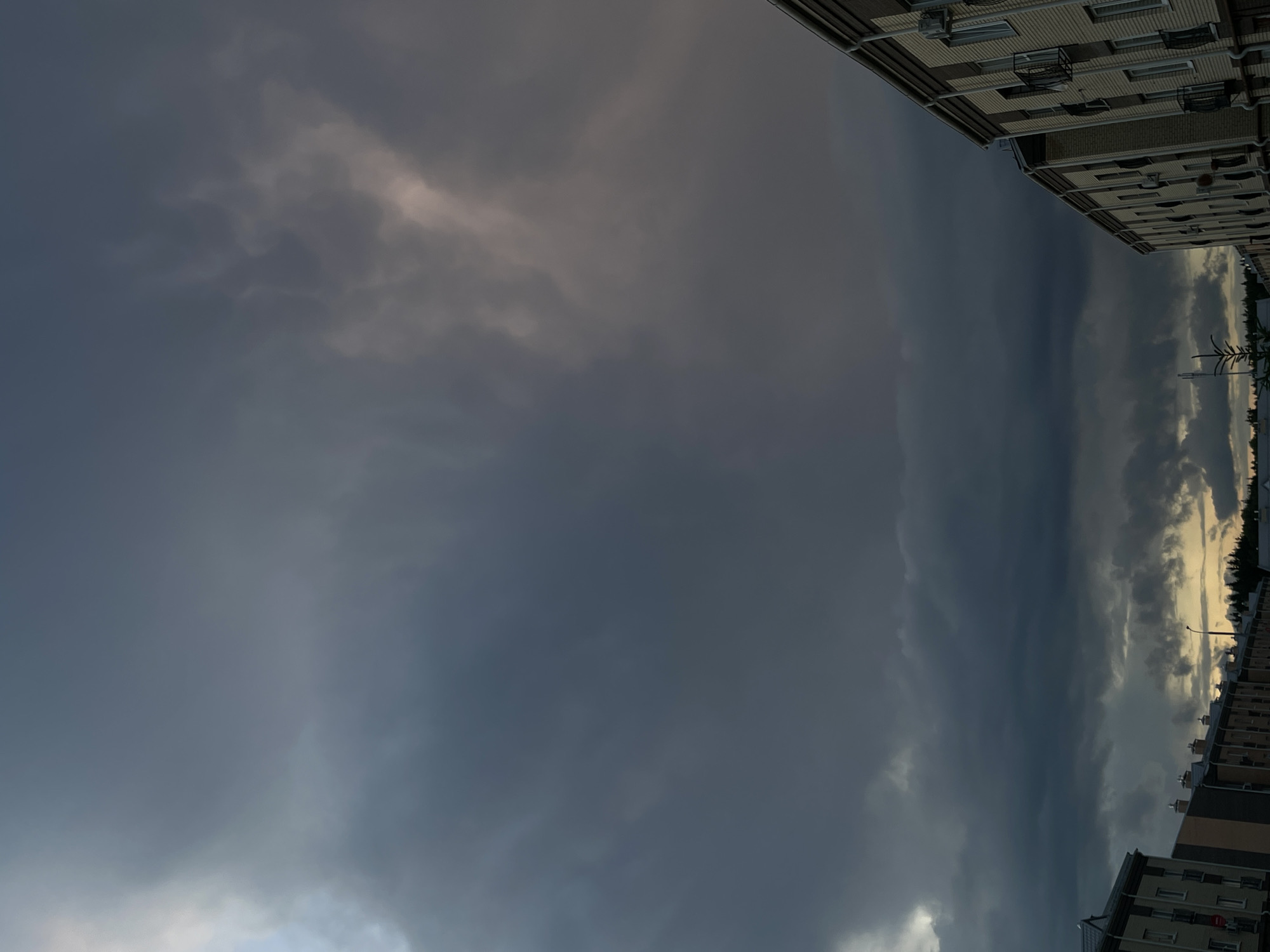 The image shows a dusky sky filled with dark, heavy clouds over a residential area in ЖК «Борисоглебское», Russia. The sky occupies the majority of the frame, with the tops of buildings visible along the bottom. The clouds appear to be moving, with some lighter patches suggesting the presence of the setting sun or breaks in the cloud cover. The buildings are multi-story structures with visible windows and balconies. The overall mood is somber, with the dark clouds creating a sense of impending weather.