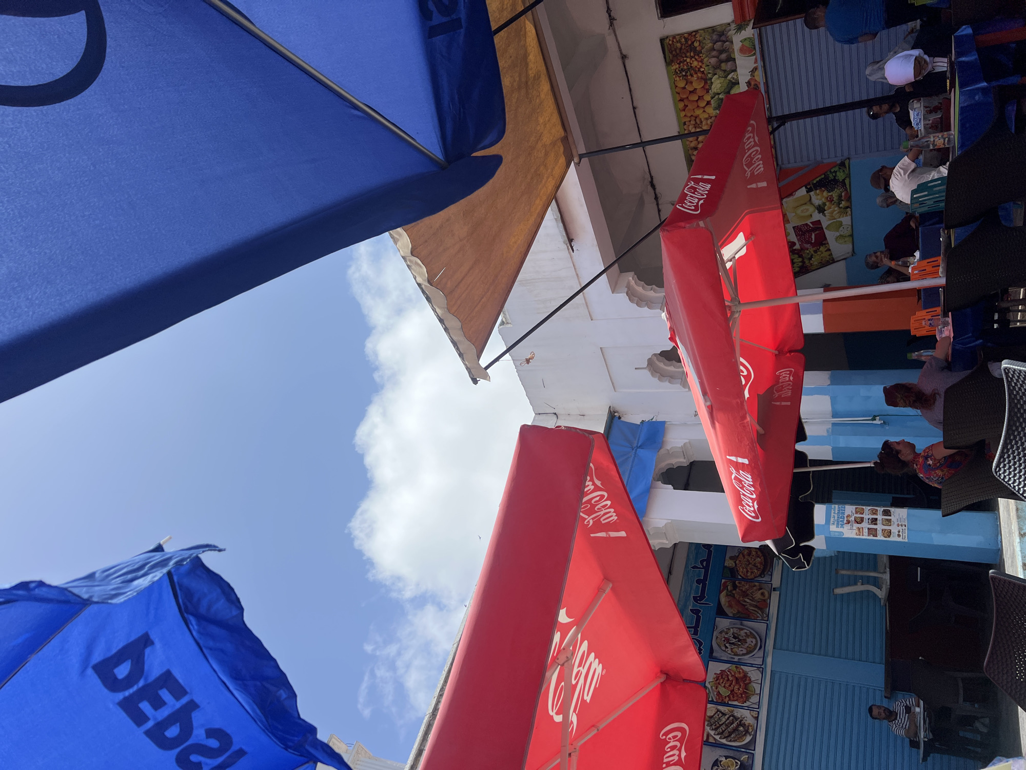 This image captures an outdoor cafe or restaurant scene from a low-angle perspective, looking upwards towards a bright blue sky with scattered white clouds. The setting appears to be a bustling street or alleyway in an urban area, consistent with Casablanca, Morocco.
Prominently featured are several large outdoor umbrellas providing shade. Two blue umbrellas are visible, one in the upper left bearing a partial circular logo and another in the lower left with the mirrored text "DEBA2". Multiple red Coca-Cola branded umbrellas are also present, especially on the right side of the frame, indicating a partnership or popular beverage offering.
Beneath the umbrellas, several people are seated at dark tables with matching chairs, suggesting they are patrons of the establishment. Their faces and activities are partially obscured by the umbrellas and furniture, but they appear to be relaxing, eating, or socializing. One individual wears a white top and a brown head covering, another a blue shirt, and a person in a striped shirt is also visible.
The surrounding buildings feature white and light blue facades, with some traditional architectural details. Brown or tan awnings extend from some of these structures. Posters and signs adorn the walls, displaying vibrant images of food items such as seafood dishes, fruits, and vegetables. Some Arabic text is visible on these signs, including the word "مدريد" (Madrid) on one menu poster, reinforcing the Moroccan location. A rolled-down metal shutter suggests a closed storefront or part of the building. The bright daylight and clear sky indicate a sunny day.
