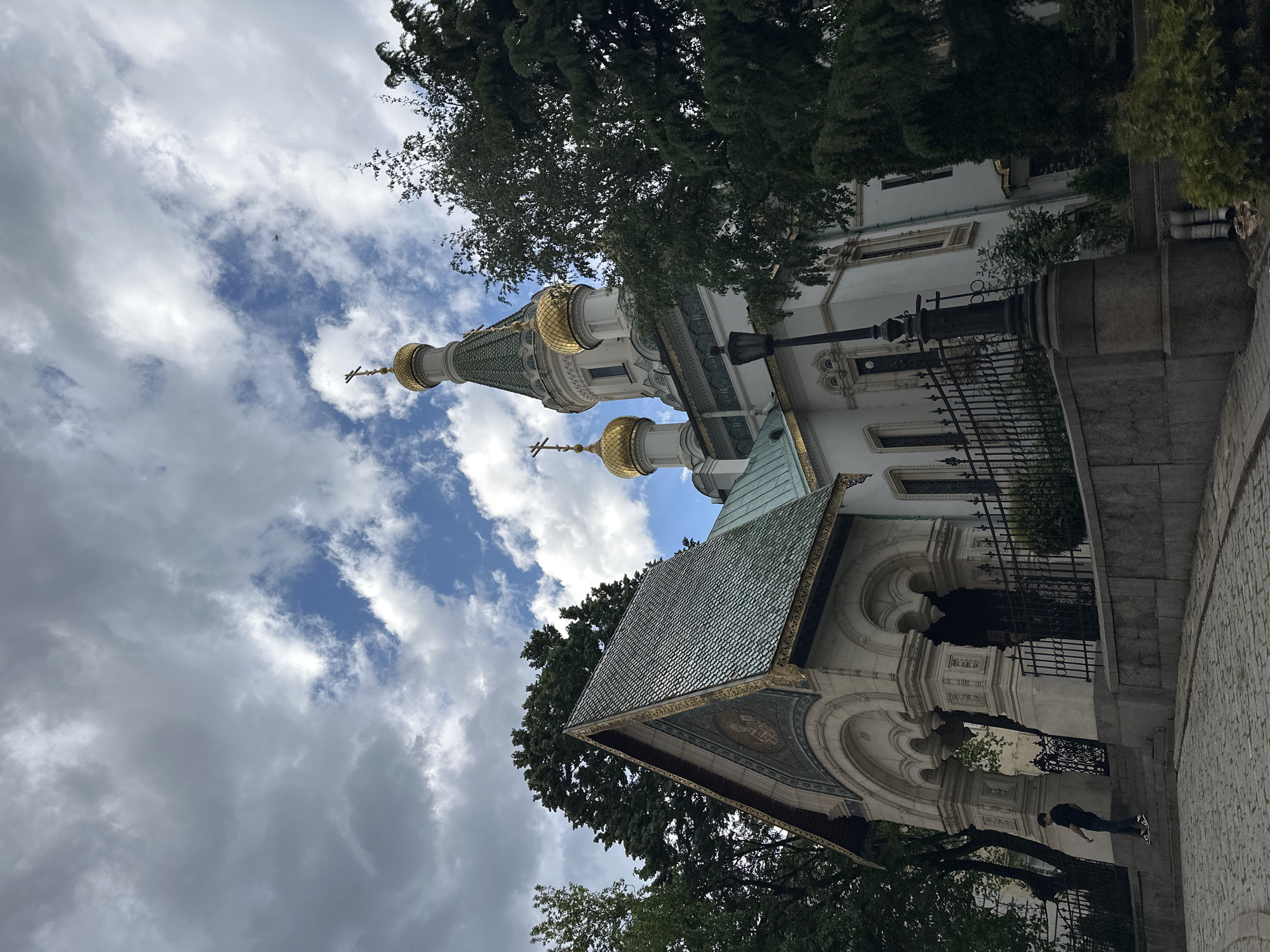 The image shows the Russian Church St. Nicholas the Wonderworker in Sofia, Bulgaria, from a low-angle perspective. The church's distinctive golden domes are visible against a cloudy sky, with some tree branches framing the architecture. The building is primarily white, with green tiling on some roofs. A black wrought-iron fence borders the church. A person is partially visible near the bottom right corner of the image. The ground is paved with cobblestones. The overall scene conveys a sense of historical architecture under a somewhat overcast sky.