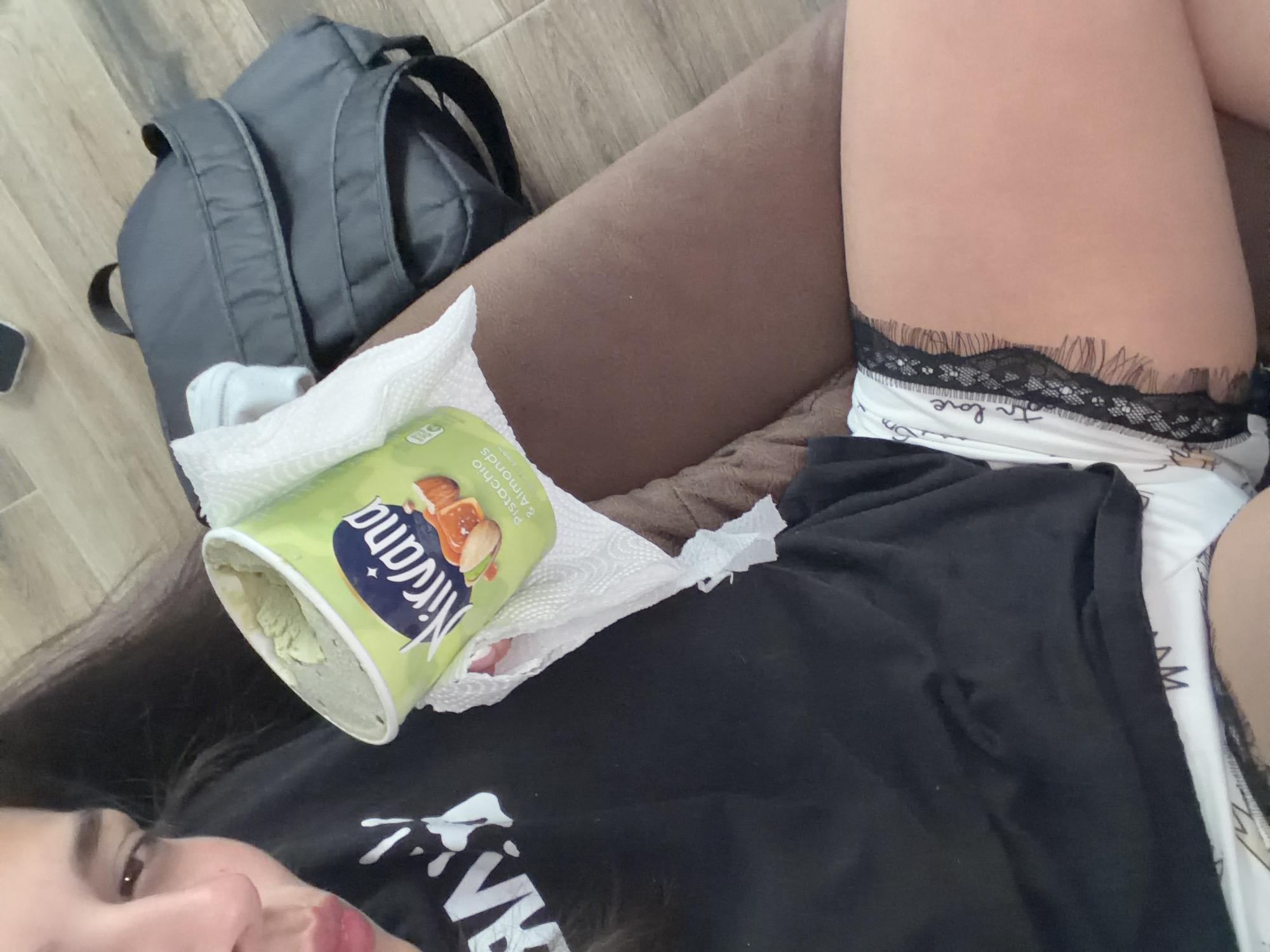 A woman is lying on a brown couch. A green container of Nirvana pistachio and almond ice cream rests on a white napkin. A gray backpack is adjacent to the couch on a light-colored floor. The woman is wearing a black shirt and white underwear with black lace trim. The location is unspecified; there is no evidence to suggest this is Gabrovo, Bulgaria.