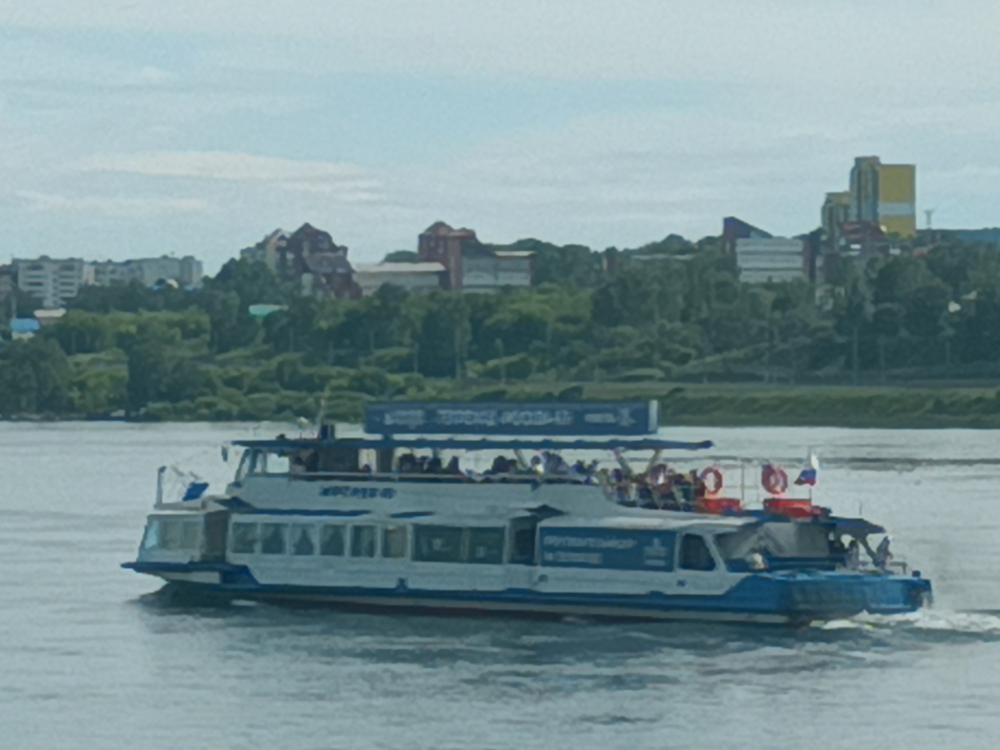 A panoramic, slightly blurry, daylight shot captures a passenger boat navigating a wide, calm river, likely the Angara River in Irkutsk, Russia. The boat, painted white with blue trim, features a covered main deck with numerous passengers visible. A second, open-air deck on top is also populated with people, some waving. The boat has a Russian flag flying from its stern. The background shows a lush green shoreline with trees and several buildings, including modern structures and older, more ornate architecture, suggesting a city setting. The sky is overcast with soft clouds.