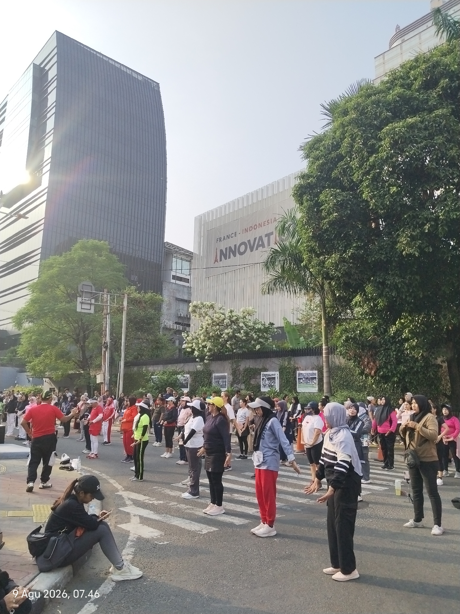 The image captures a group of people engaged in what appears to be an outdoor exercise class or a community gathering. They are spread across a paved street, with some standing and others in motion, suggesting a choreographed activity. The participants are diverse in age and attire, with many wearing athletic wear. The scene is set in an urban environment, characterized by modern buildings, including a tall glass skyscraper on the left and a building with signage reading "FRANCE-INDONESIA INNOVAT" in the background. Lush green trees are present, providing a contrast to the architectural structures. The lighting suggests it is daytime, possibly morning or late afternoon. The image also includes a young woman sitting on a curb in the foreground, looking at her phone, detached from the main activity. The timestamp "9 Agu 2026, 07.46" is visible in the lower left corner, indicating the date and time the photograph was taken. The location is identified as Central Jakarta, Indonesia.