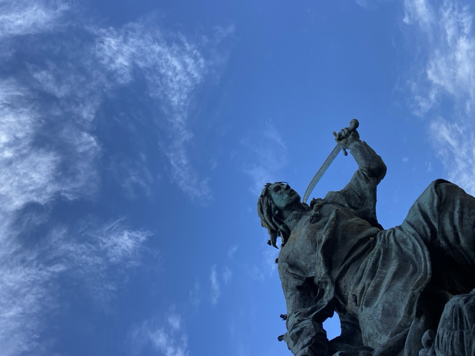 A low-angle shot captures a bronze statue against a vibrant blue sky dotted with wispy clouds. The statue appears to be of a man with flowing hair, holding a curved sword aloft. The figure is positioned in the lower right corner, while the cloudy sky dominates the upper left, creating a strong contrast. The statue’s detailed texture suggests weathering.